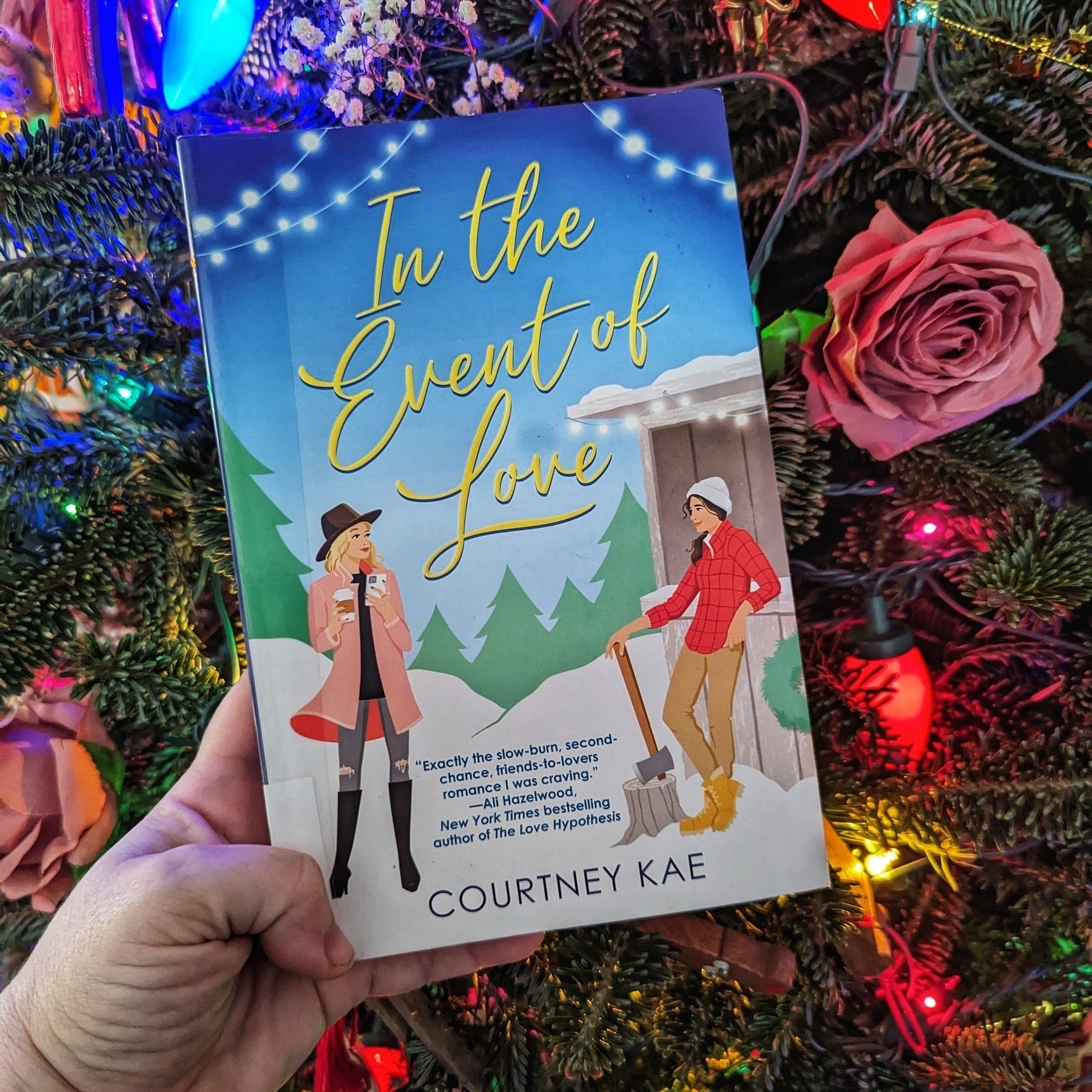 A hand holding a book titled “In the Event of Love” by Courtney Kae in front of a colorful Christmas tree, with a rose and lights visible in the background. The book is a romantic novel set in a winter wonderland.