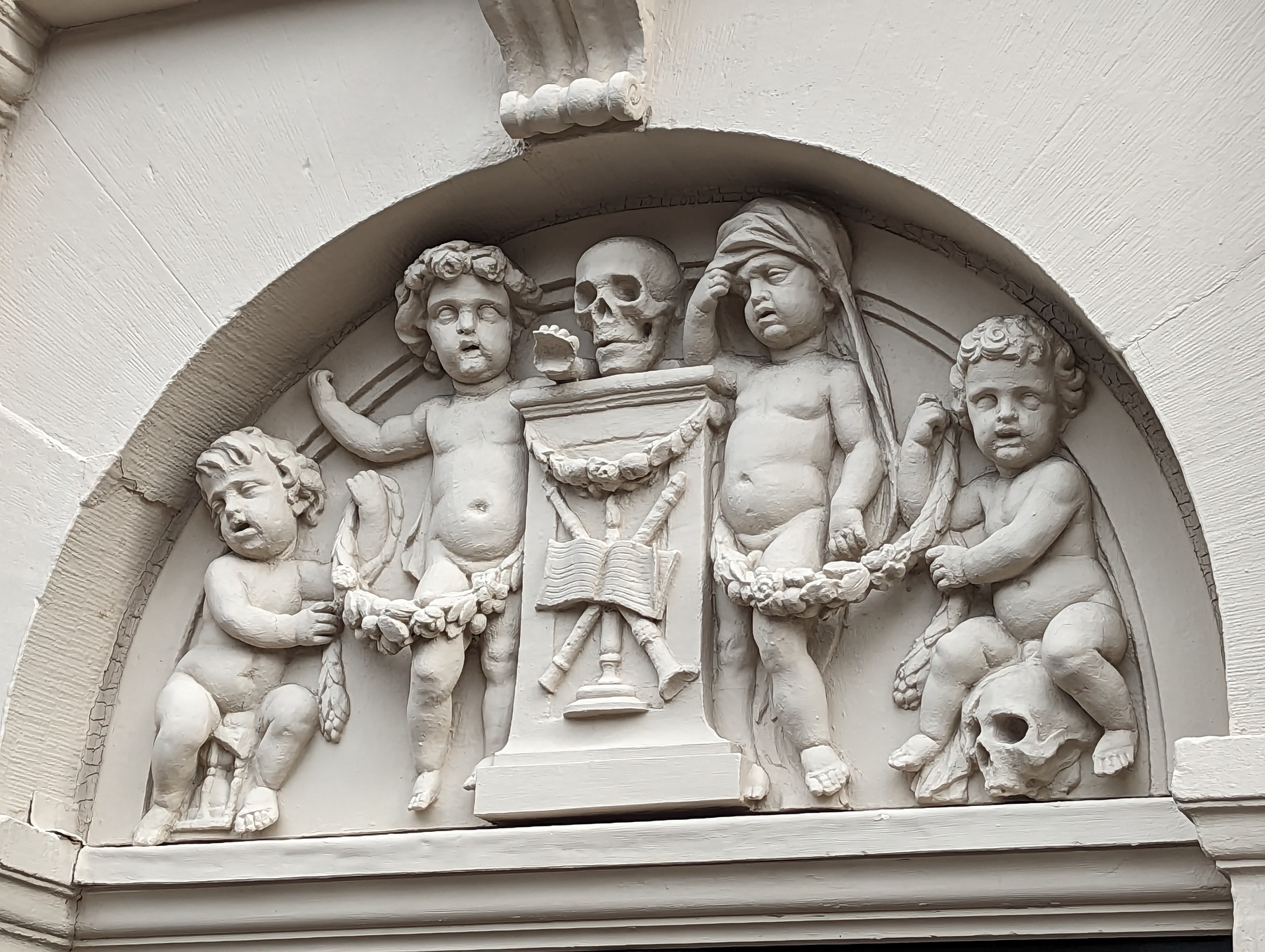 A stone relief. A skull sits on a pedestal. The pedestal is adorned with woodwind instruments and sheet music. Four babies surround the pedestal, two on each side. The two closest to the pedestal stand. The baby on the far left sits on an hourglass. The baby on the far right sits on a skull.