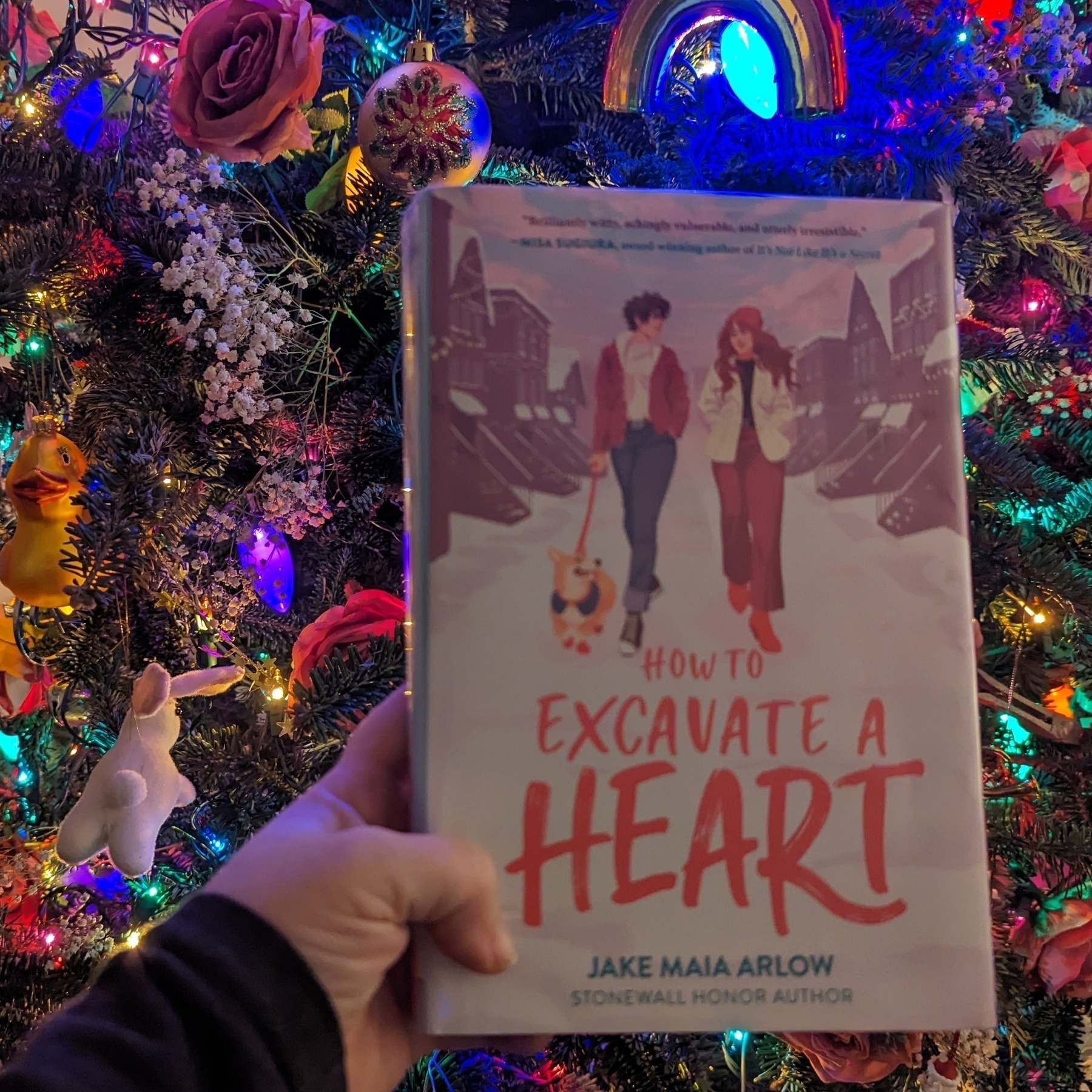 A hand holding a book titled 'How to Excavate a Heart' by Jake Maia Arlow in front of a brightly decorated Christmas tree.