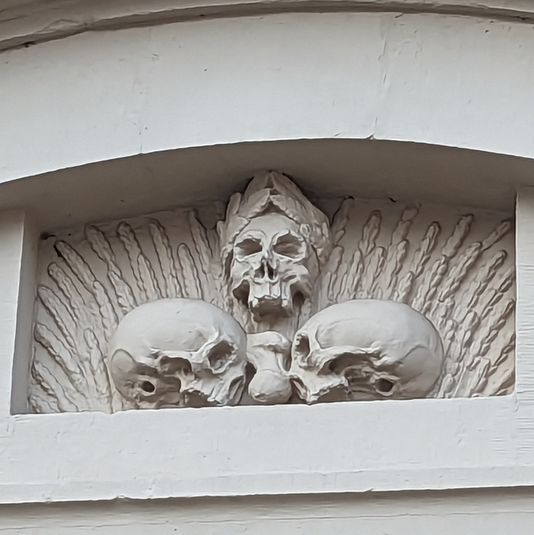 A stone relief. A skull wearing a leaf crown floats over two other skulls. The crowned skull faces forward. The other two skulls are in profile.