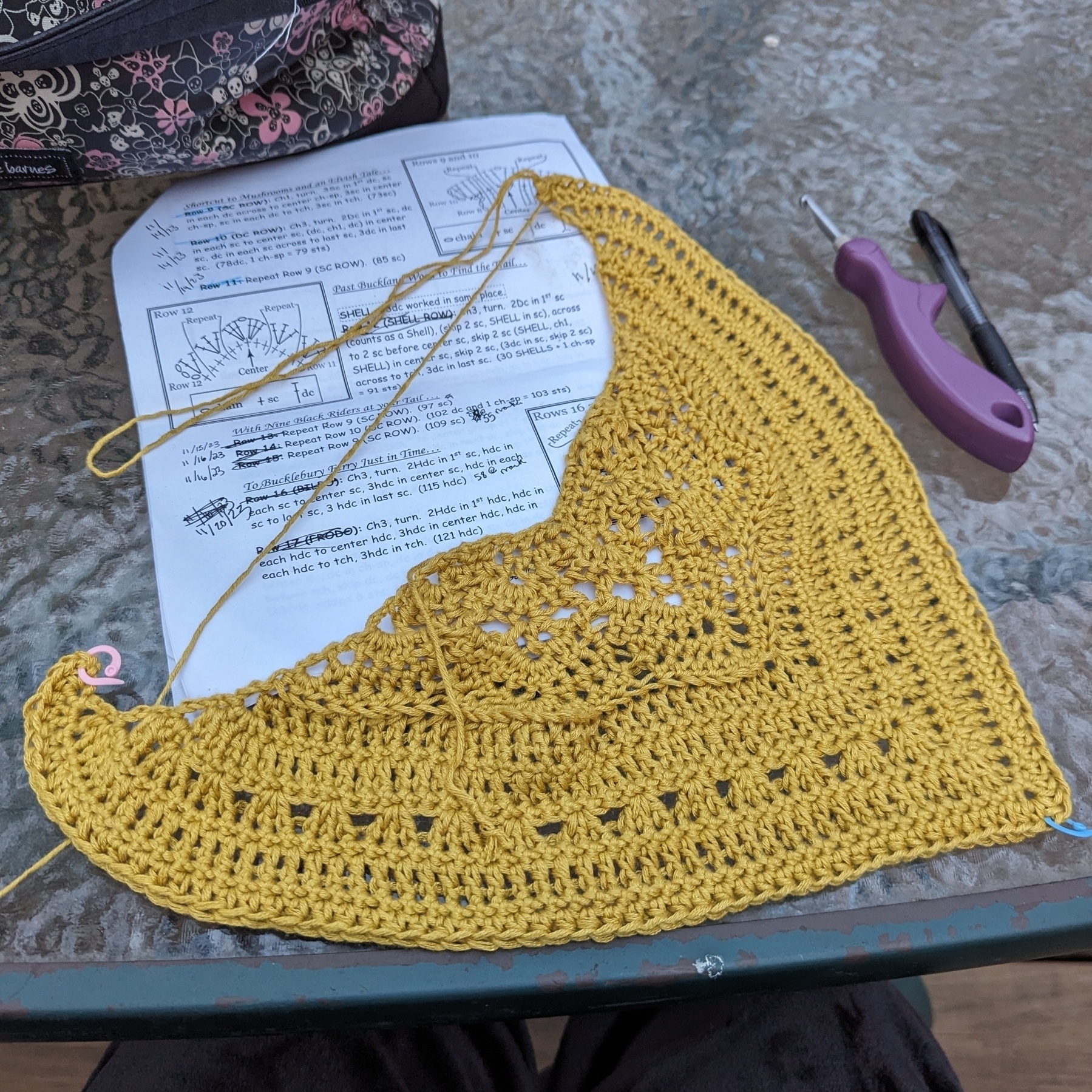 A golden-colored crocheted shawl, still in progress, sits on top of a printed pattern. To the right of it are a crochet hook and black pen.