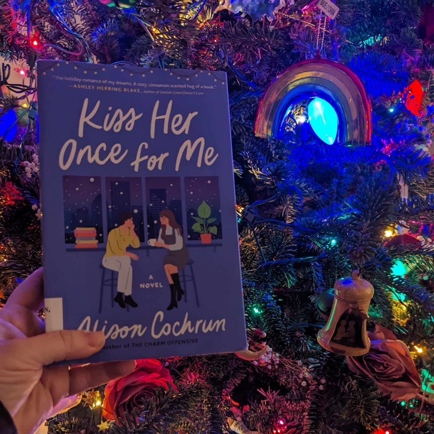 A hand holding a book titled 'Kiss Her Once for Me' by Alison Cochrum in front of a brightly decorated Christmas tree with colorful lights and ornaments.