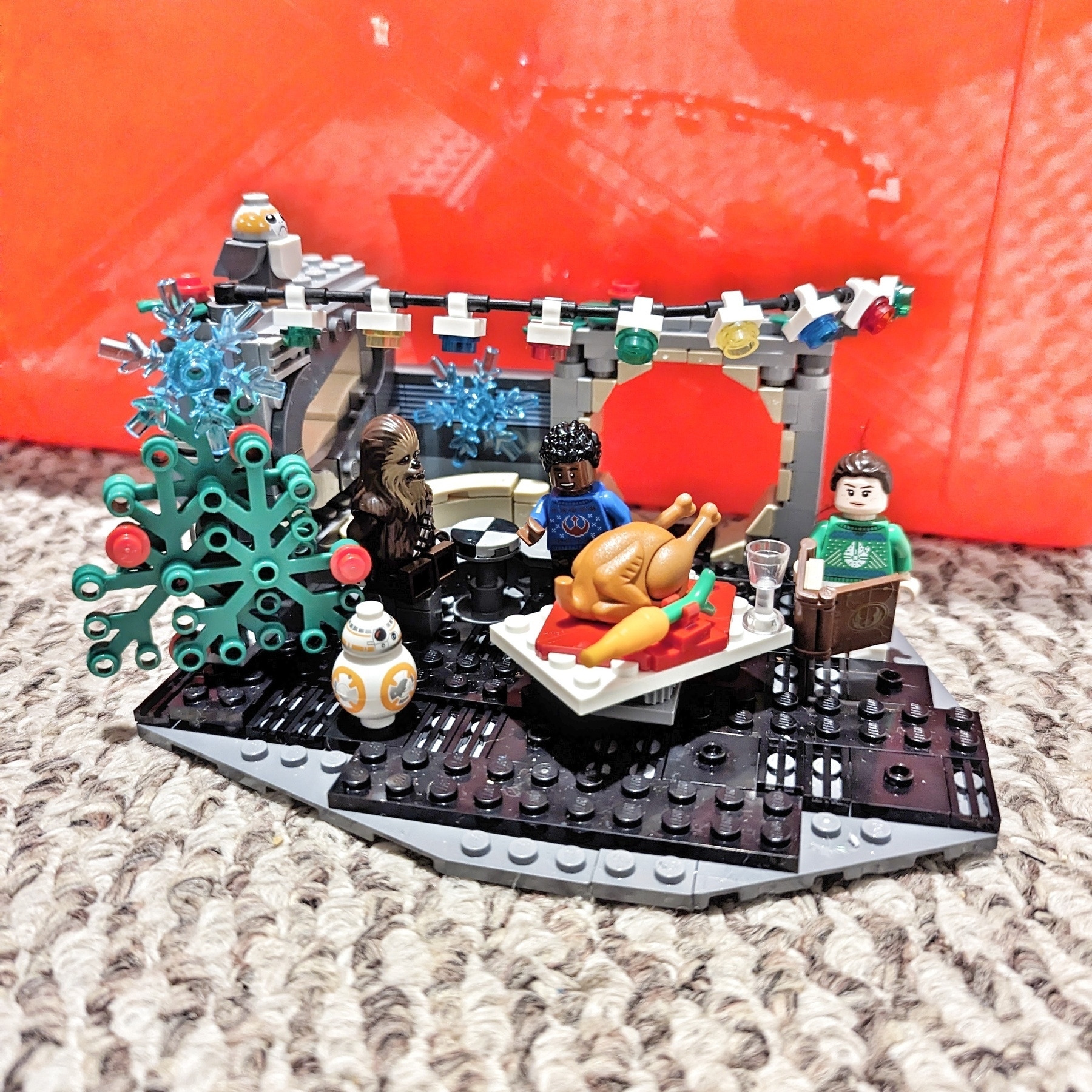 A LEGO set. In the Millennium Falcon, Chewbacca, Rey, Finn, BB-8, and a Porg celebrate Life Day with a tree, hanging lights, and some kind of cooked fowl with a carrot.
