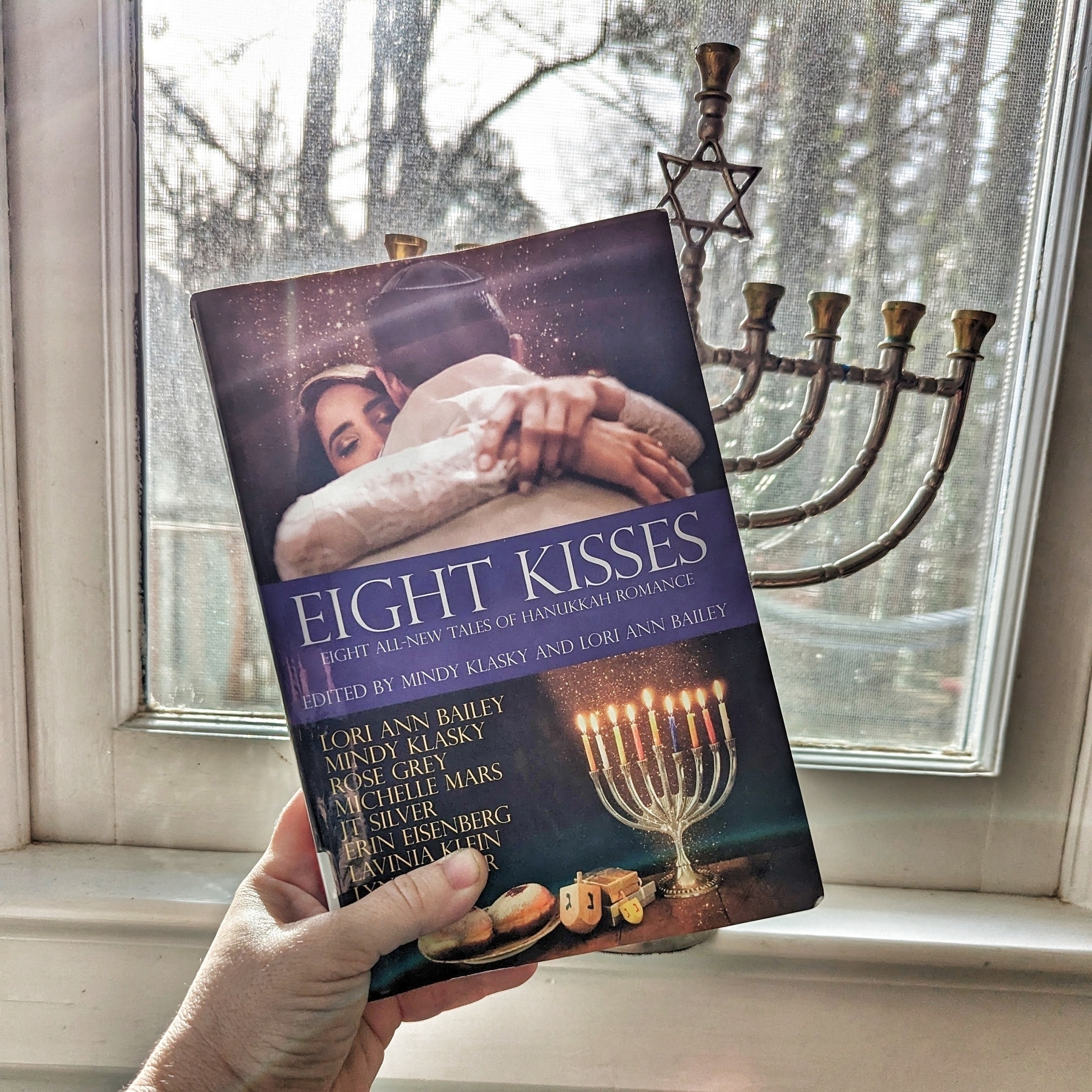 A hand holding the book “Eight Kisses” in front of a window with a menorah on the sill, showcasing a cover featuring two people embracing and a lit menorah. The book is a collection of eight stories of Hanukkah romance by various authors.