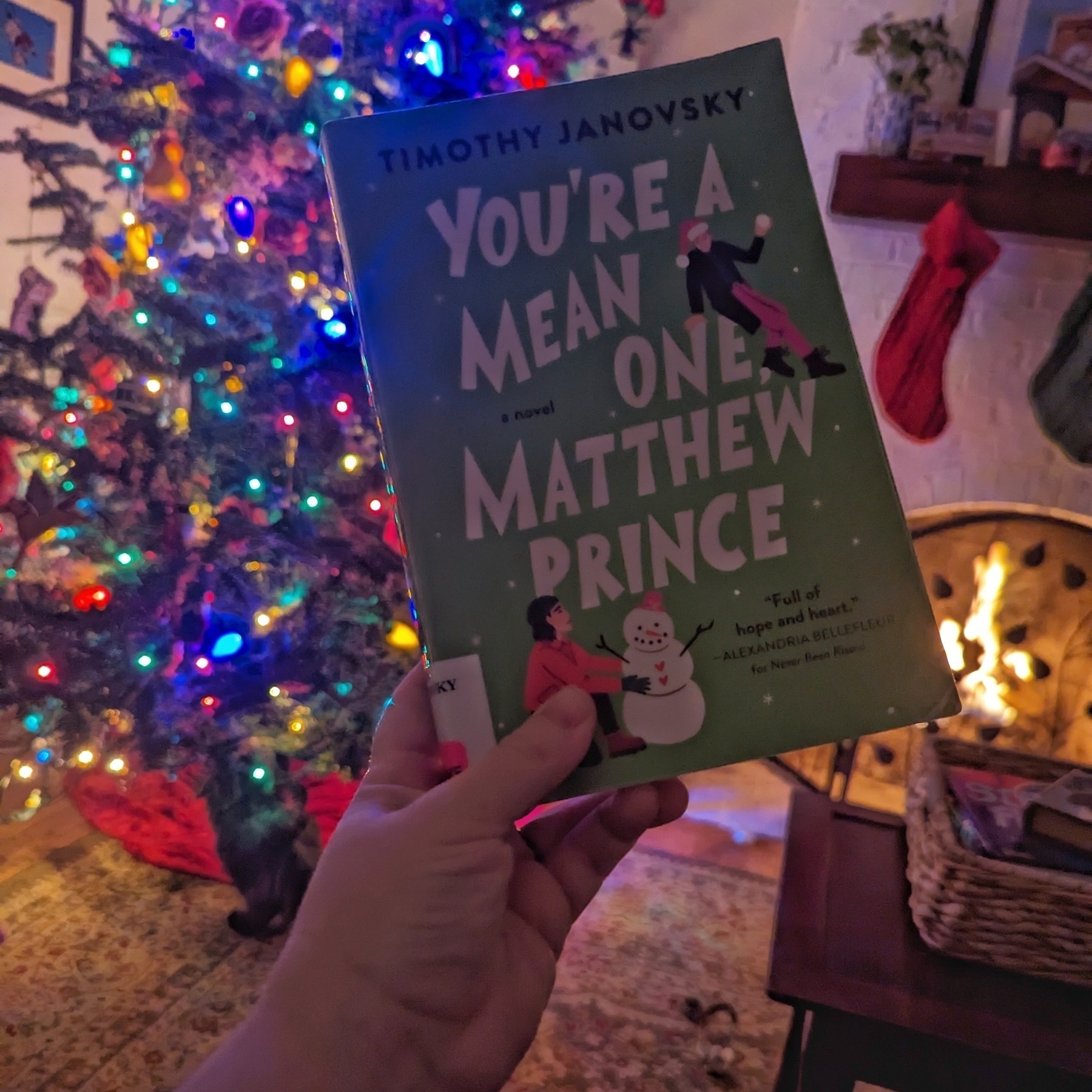 A hand holding a book titled YOU'RE A MEAN ONE MATTHEW PRINCE by TIMOTHY JANOVSKY in front of a decorated Christmas tree and a lit fireplace.