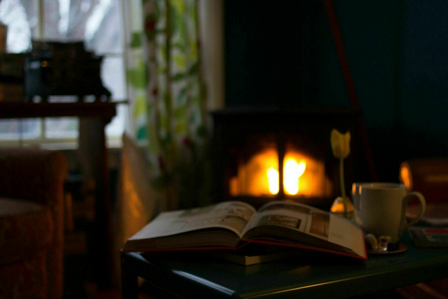 A book is open on a table. A fire in a fireplace is in the background.