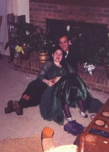 A young man sits in front of a fireplace, his arms wrapped around a young woman who sits with him.