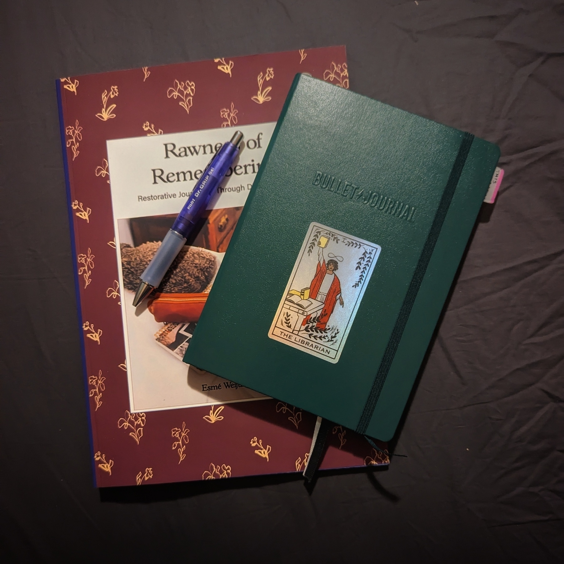 Two books and a pen on a dark surface. One book is titled “Rawness of Remembering: Restorative Journaling through Difficult Times” and has a purple cover with golden designs. The other book is a green bullet journal with a Librarian Tarot card sticker. A purple pen rests on the purple book.