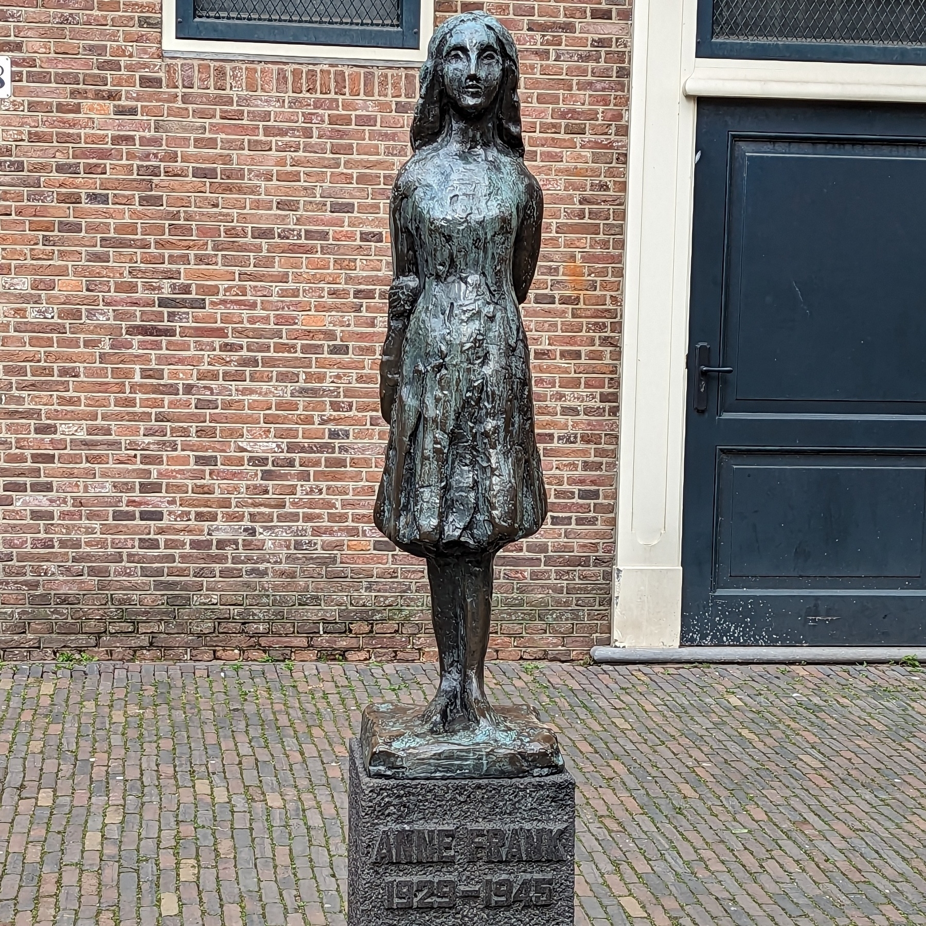 Auto-generated description: A bronze statue of Anne Frank stands in front of a brick wall on a cobblestone walkway.