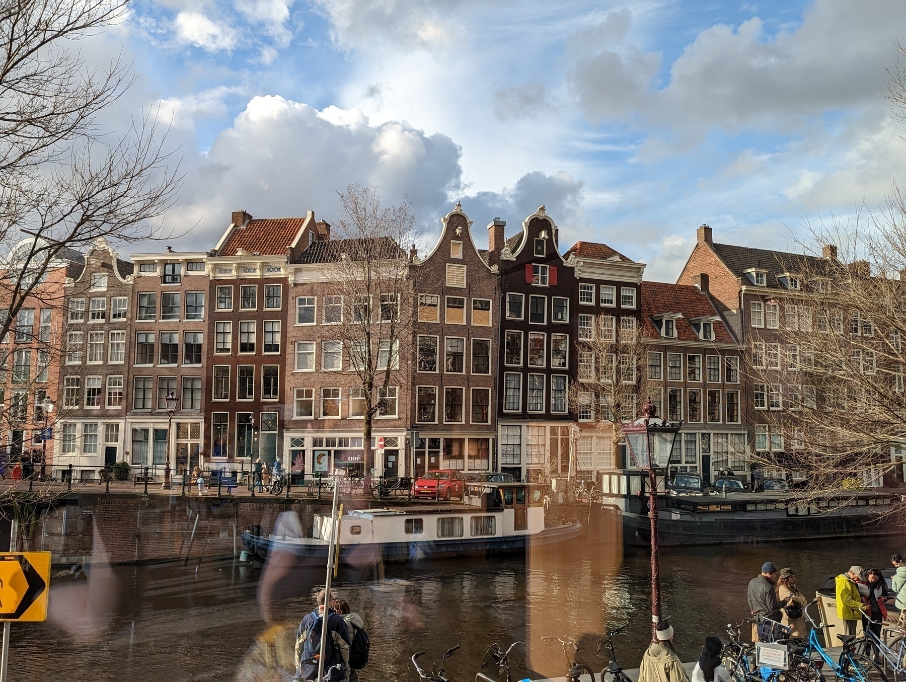 A picturesque canal scene features traditional Dutch row houses, a boat on the water, and people walking and biking nearby, with a reflection in a glass window.