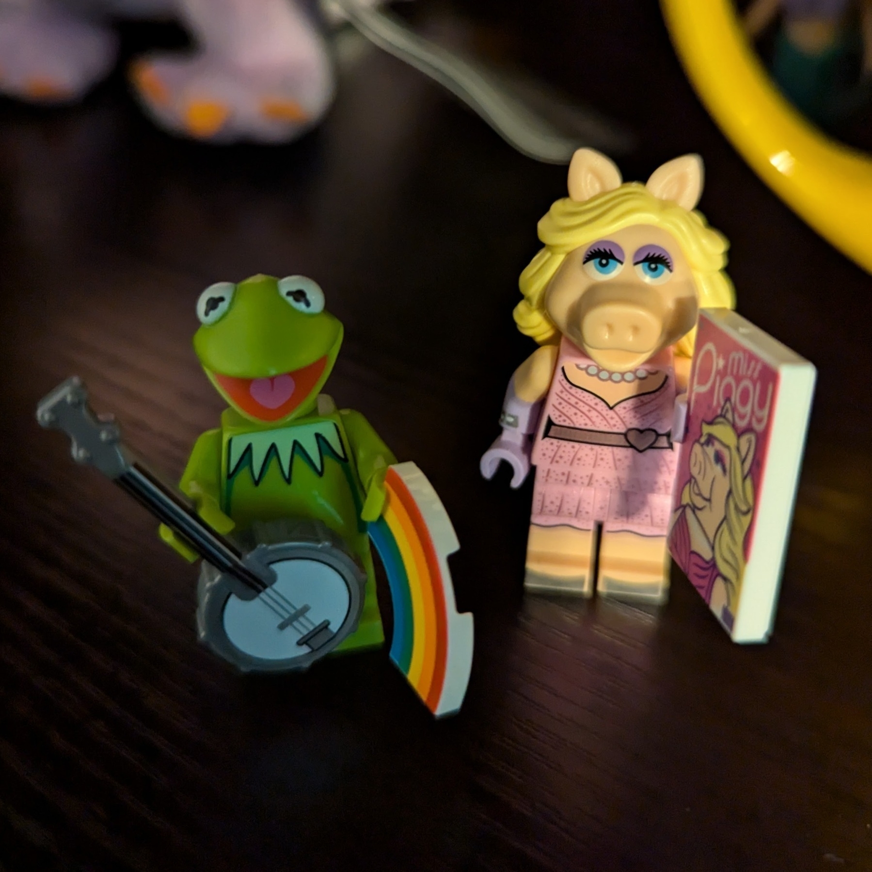 Auto-generated description: Two toy figures, one resembling a green frog with a banjo and a rainbow, and the other resembling a pig holding a book, are displayed on a dark surface.