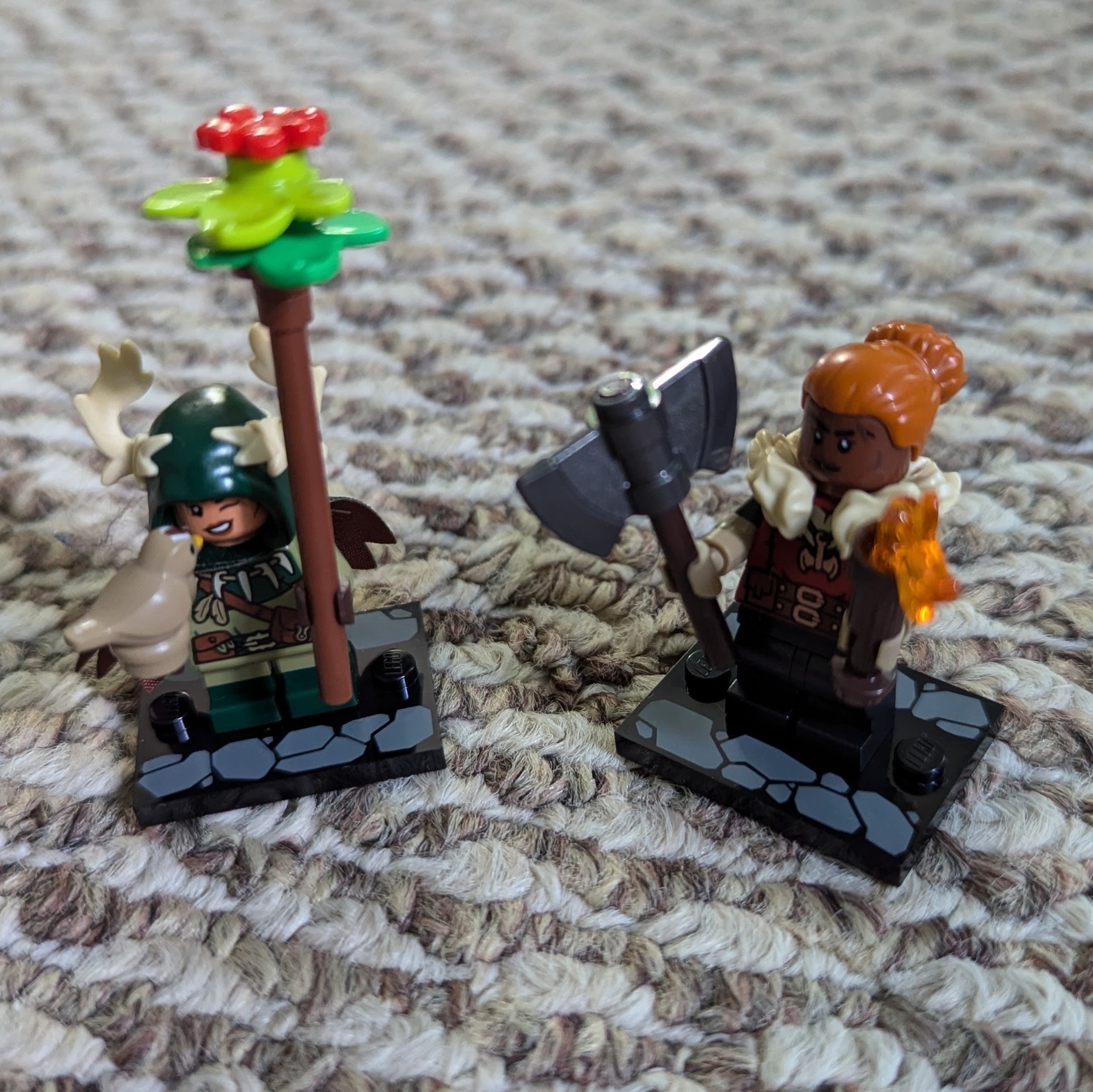 Auto-generated description: Two LEGO minifigures are standing on a carpet, one dressed in green with a staff and the other in brown with an axe and flame accessory.