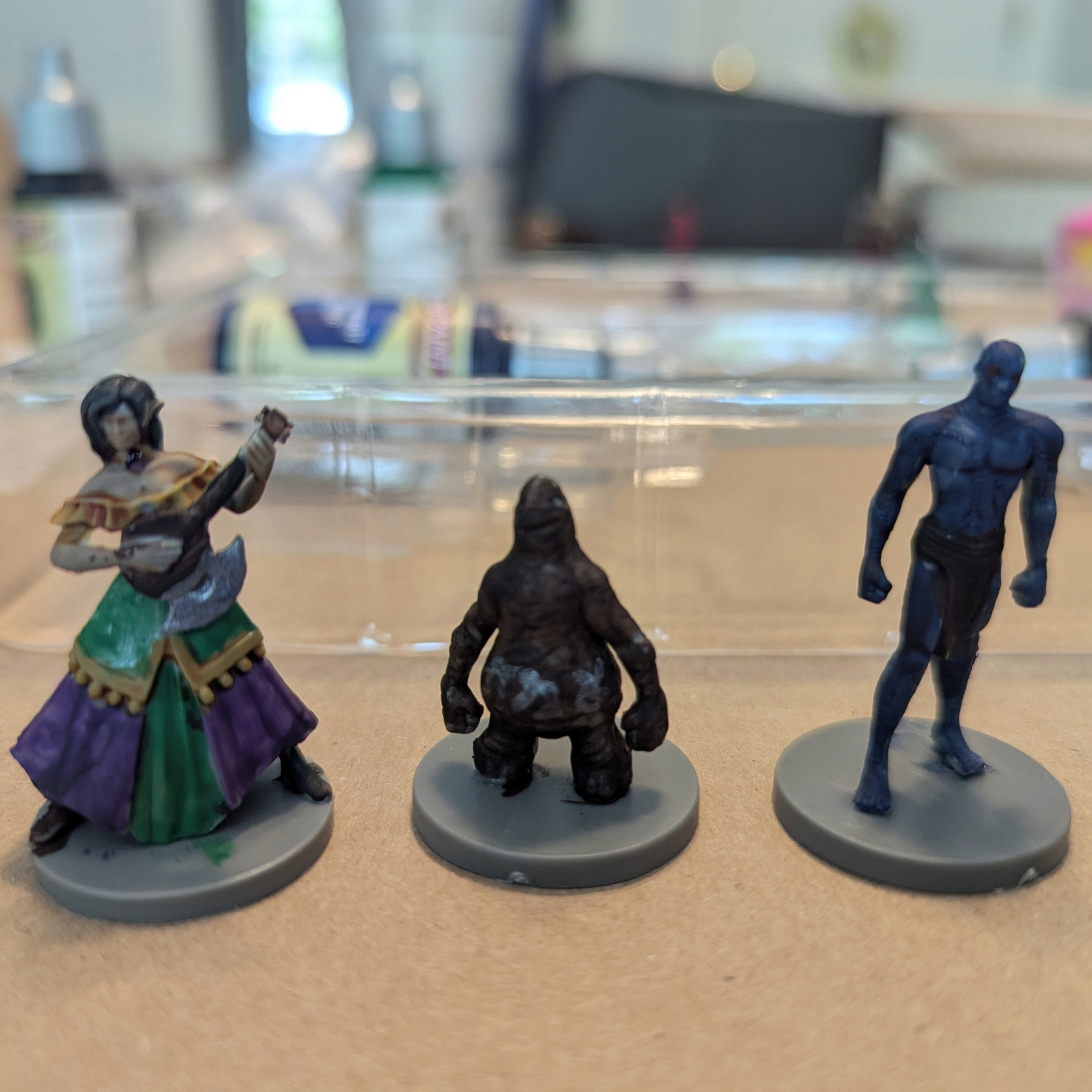 Three painted miniature figurines, including a woman playing a lute, a crouching creature, and a muscular blue humanoid, are displayed on a table.