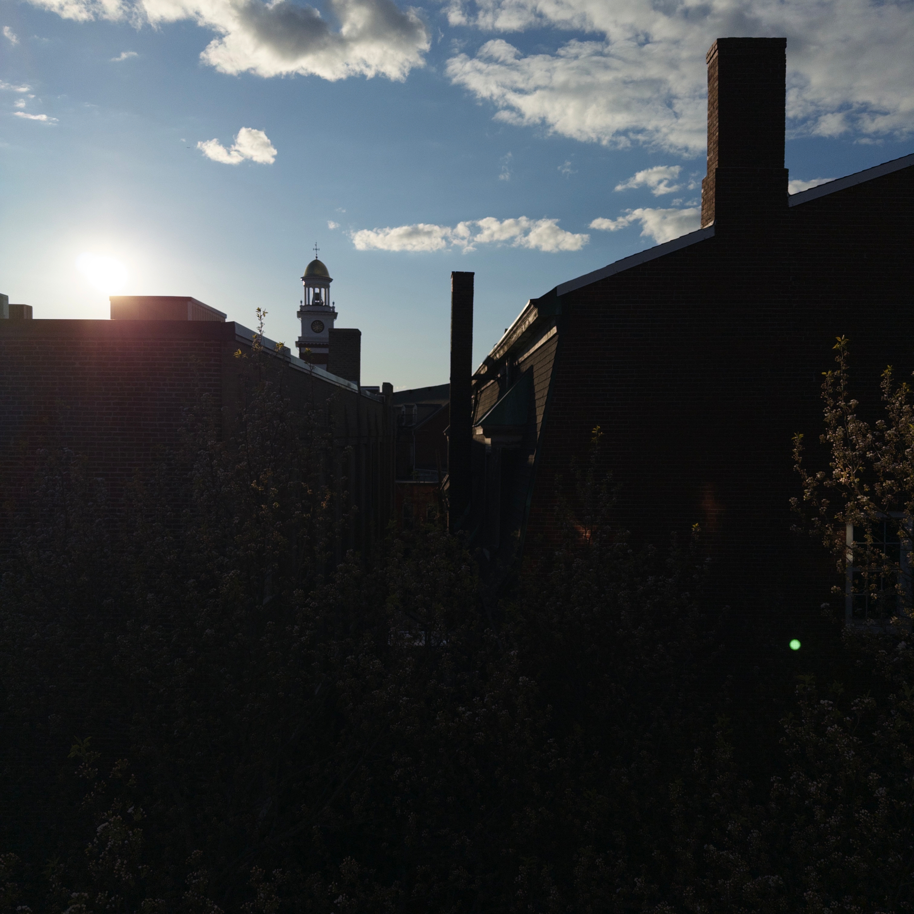 The silhouette of buildings is set against a sunset sky with clouds and a partially visible sun.