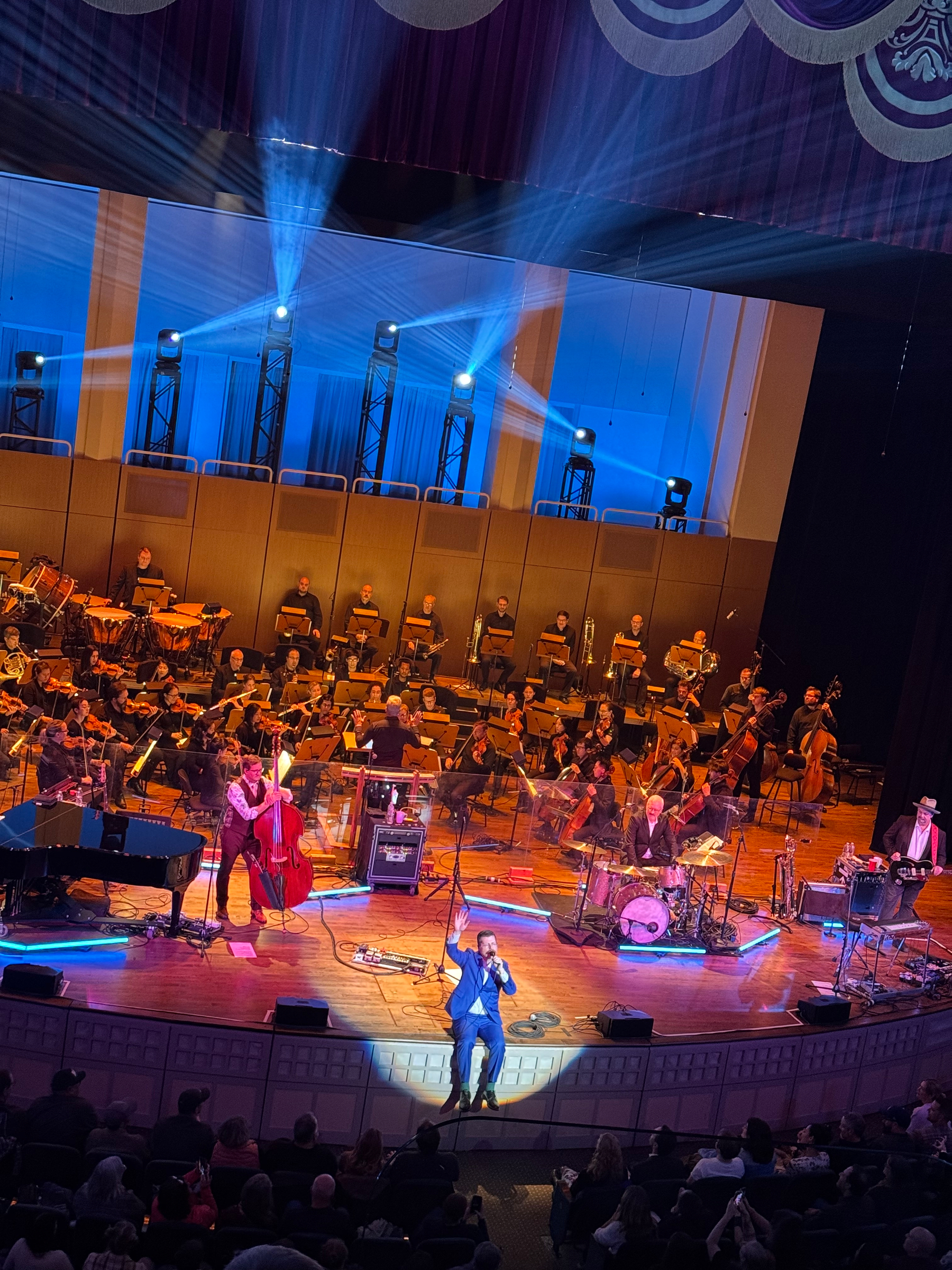 On stage at the Arlene Schnitzer Concert Hall, the Decemberists perform with the Oregon Symphony. Lead singer Colin Meloy, wearing a blue suit, sits at the very edge of the stage under a bright white spotlight, holding a microphone and gesturing with one hand toward the audience. Behind him, the orchestra fills the stage, with rows of string players, brass, and percussion lit in deep blue. The band’s instruments—a piano, upright bass, drum set, and guitars—are arranged across the front. The ornate theater interior glows with soft blue light, highlighting its carved details and gold accents. Audience members are visible in the foreground, watching attentively.