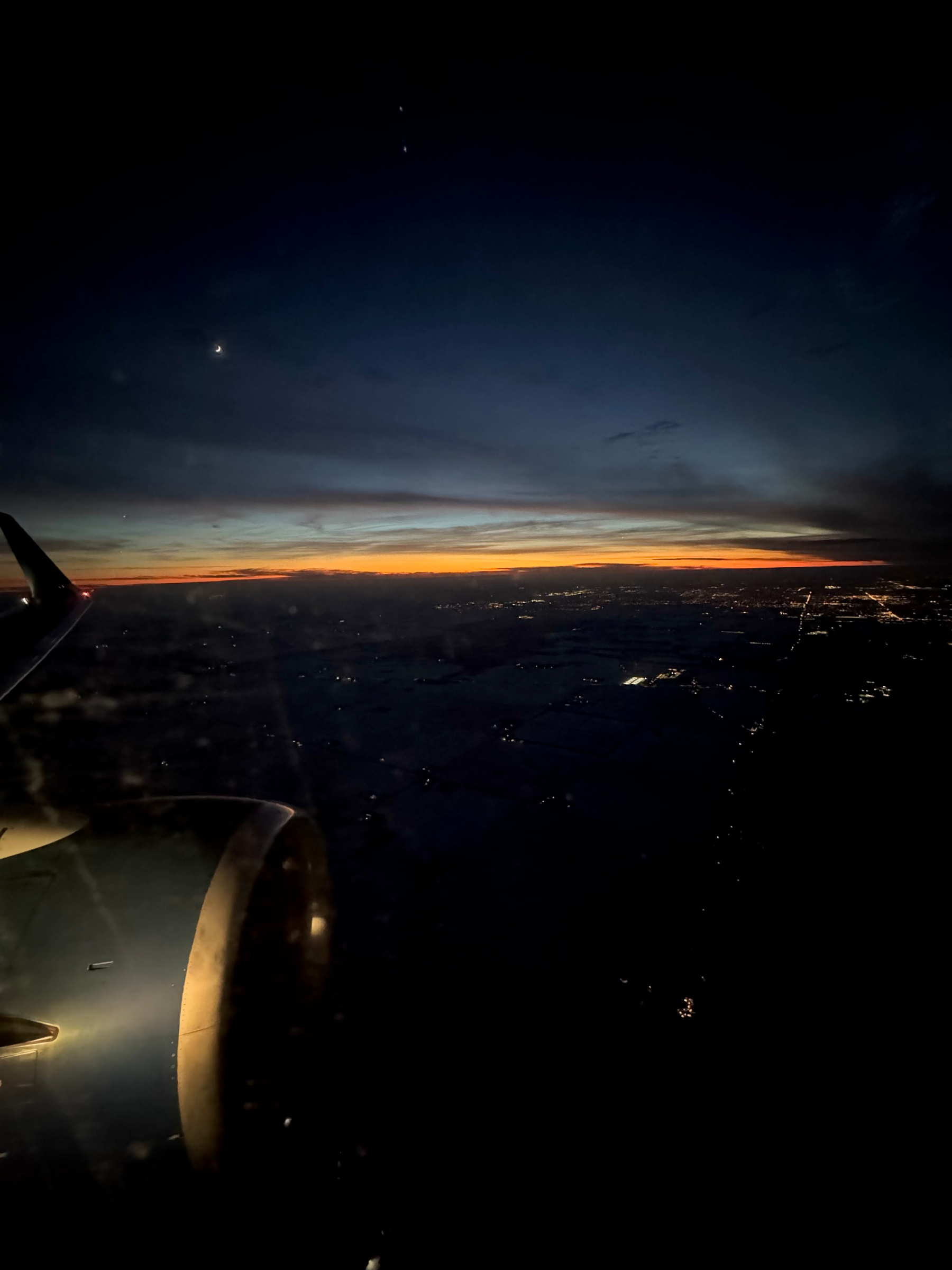 Aerial view from an airplane window at sunset, with a jet engine visible in the foreground and a thin band of orange light stretching along the horizon. The land below fades into darkness beneath layered blue-gray clouds, marking the transition from day to night.