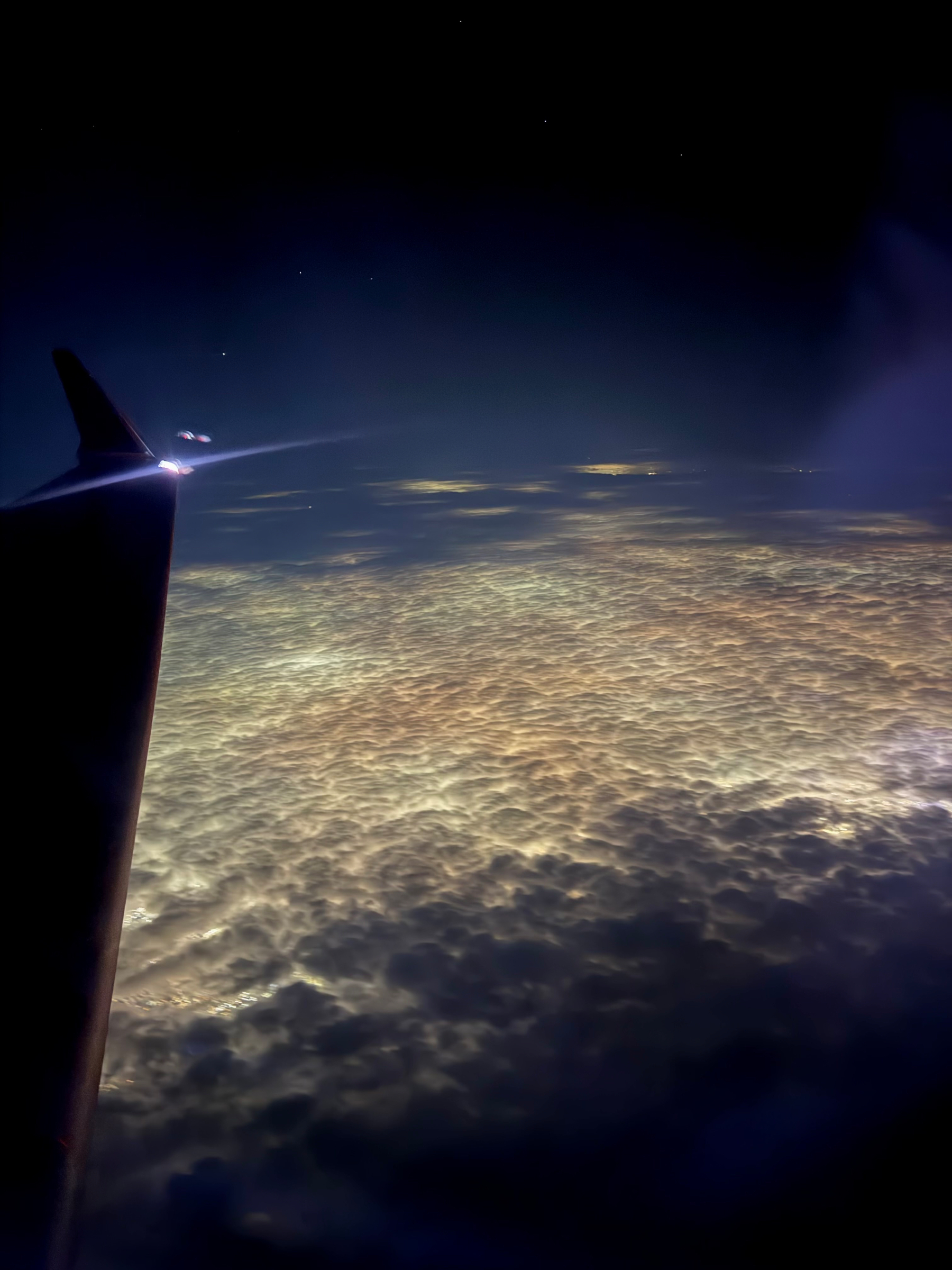 A nighttime view from an airplane window showing a wingtip light above a thick, softly textured layer of clouds. The illuminated cloud tops stretch into the distance under a dark sky, creating a quiet, abstract view of flight at cruising altitude. Probably somewhere near Rochester, NY. 