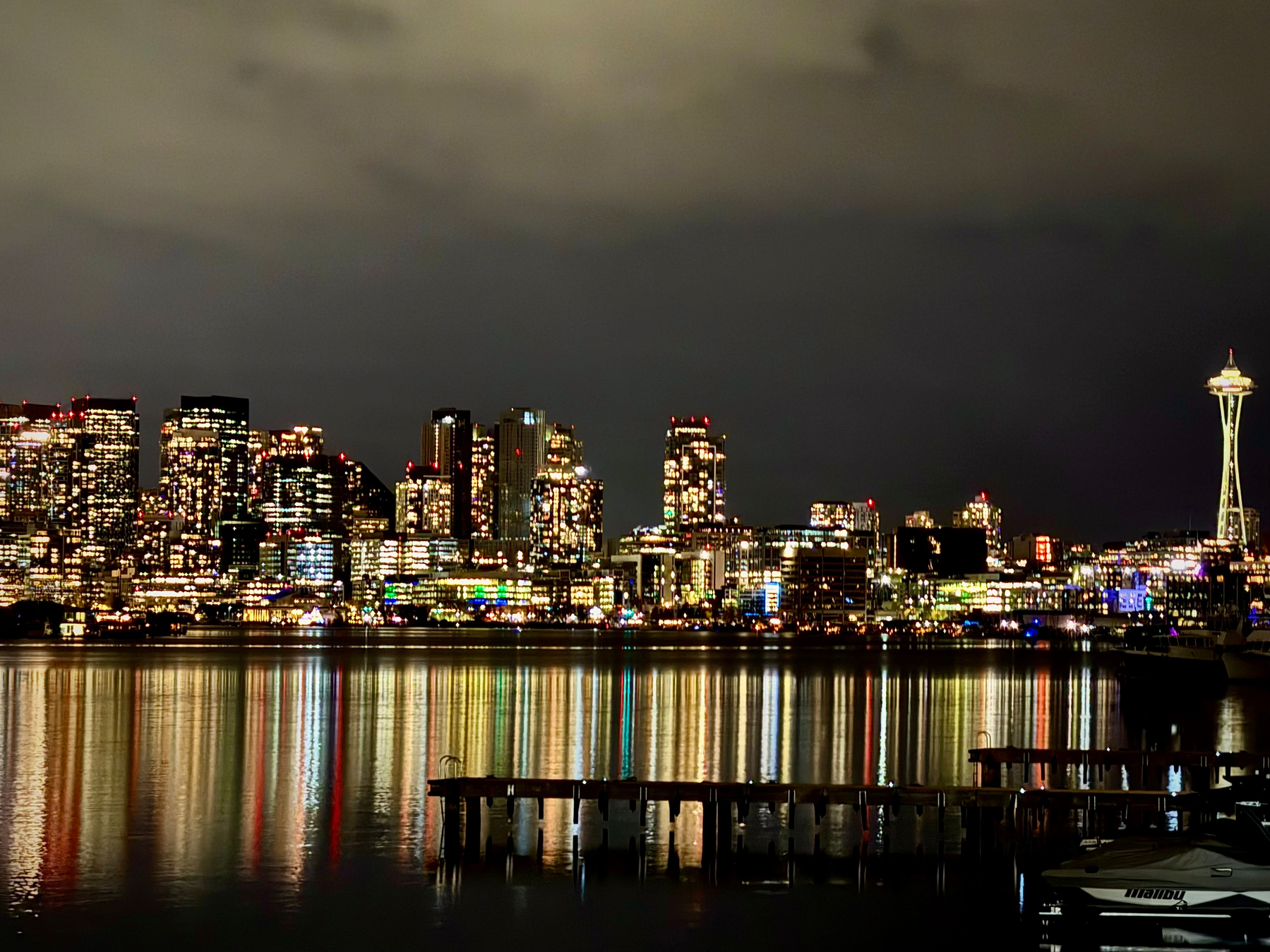 A nighttime view of the Seattle skyline across calm water, with the city’s skyscrapers glowing in warm lights and long vertical reflections stretching across the surface. The Space Needle stands on the right side of the frame, lit in soft yellow. A layer of low clouds hangs above the city, and a dark wooden dock and a small boat sit in the foreground.