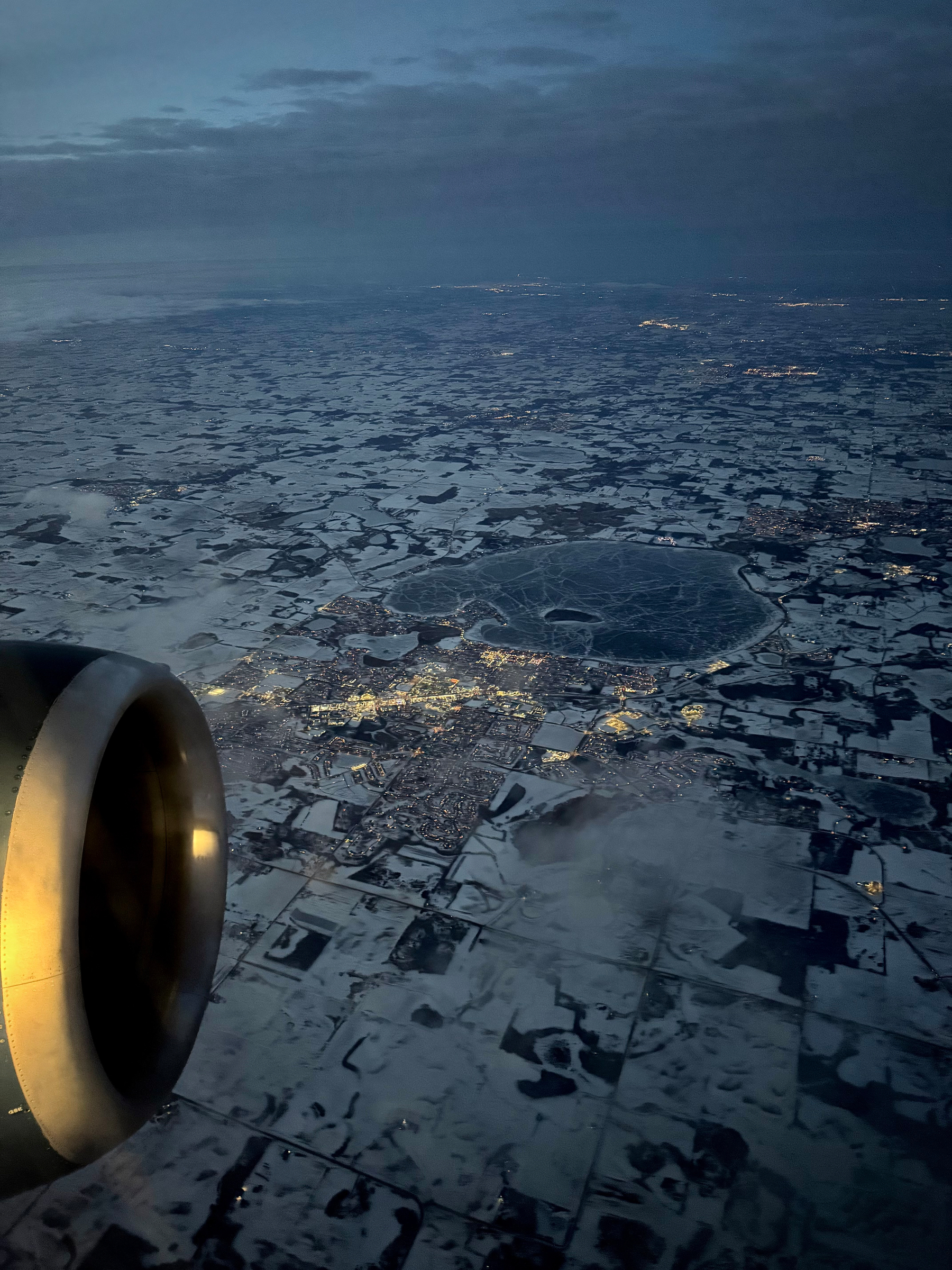A nighttime aerial view from an airplane window showing a frozen, circular lake etched with pale cracks, surrounded by snow-covered land. Warm yellow city lights cluster along the lake’s edge, contrasting with the cold blue-gray ice and darkness below. Taken somewhere in South Dakota or Minnesota. 