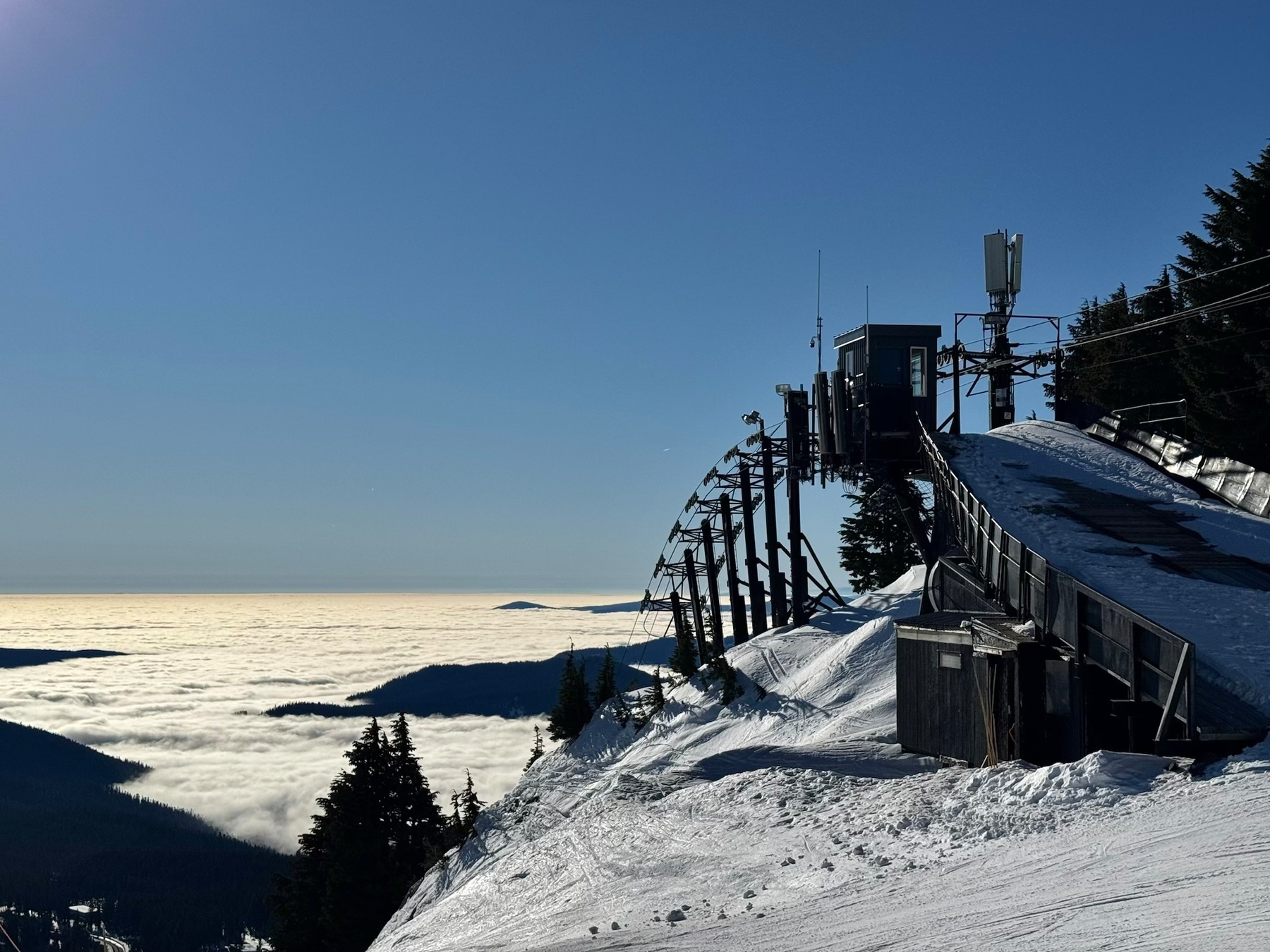 A steel ski-lift structure curves diagonally down a snowy ridgeline from the upper right toward the center of the image, with dark metal beams, platforms, cables, and small antennas silhouetted against the sky. The snow-covered slope beneath the structure shows tracks and wind-sculpted texture. Below the ridge, a vast blanket of white clouds fills the valleys, with dark forested hills protruding like islands. The sky above is a smooth gradient of clear blue. The composition emphasizes the contrast between rigid industrial geometry and the soft, expansive cloud layer below.