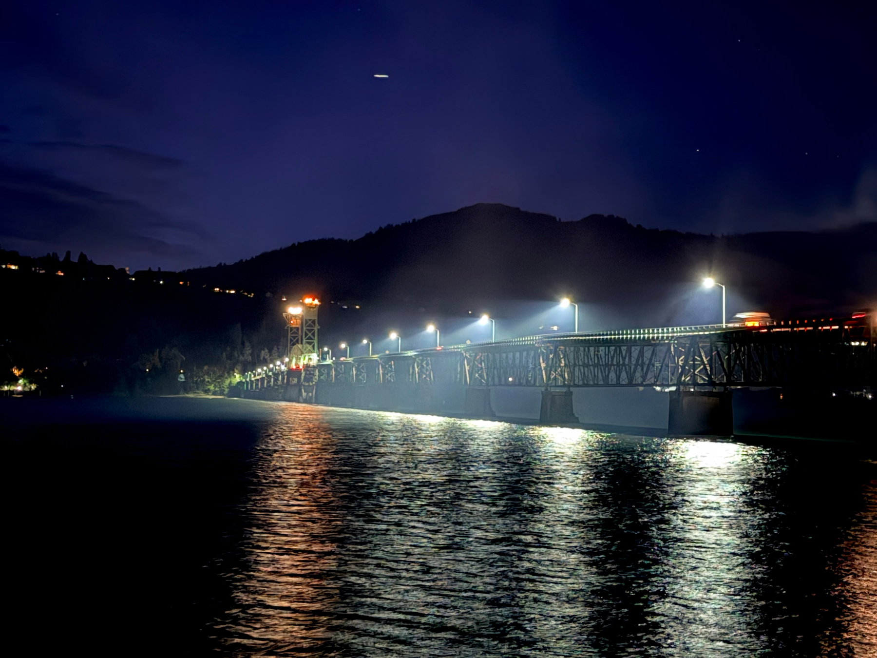 The Hood River Bridge glows with evenly spaced lights against a deep blue night sky. Reflections shimmer across the river while the surrounding hills fade into silhouette.