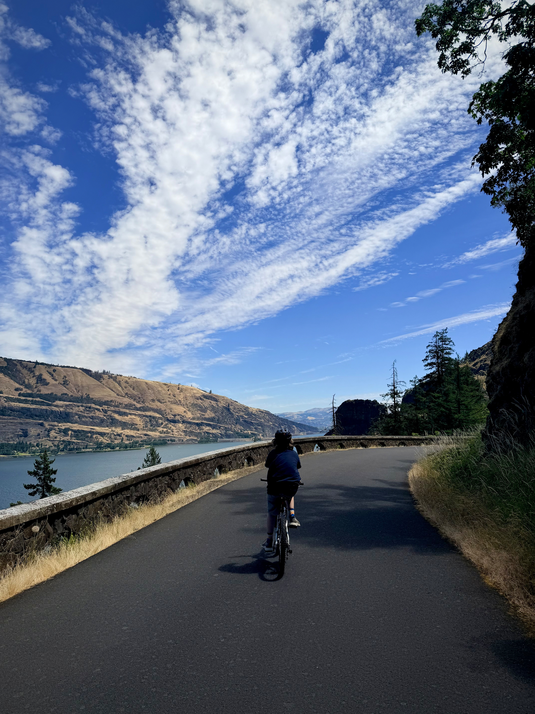 A child rides a bicycle along a smooth paved trail high above the Columbia River Gorge. Dramatic white clouds sweep across a deep blue sky, with golden hills and the river stretching into the distance.