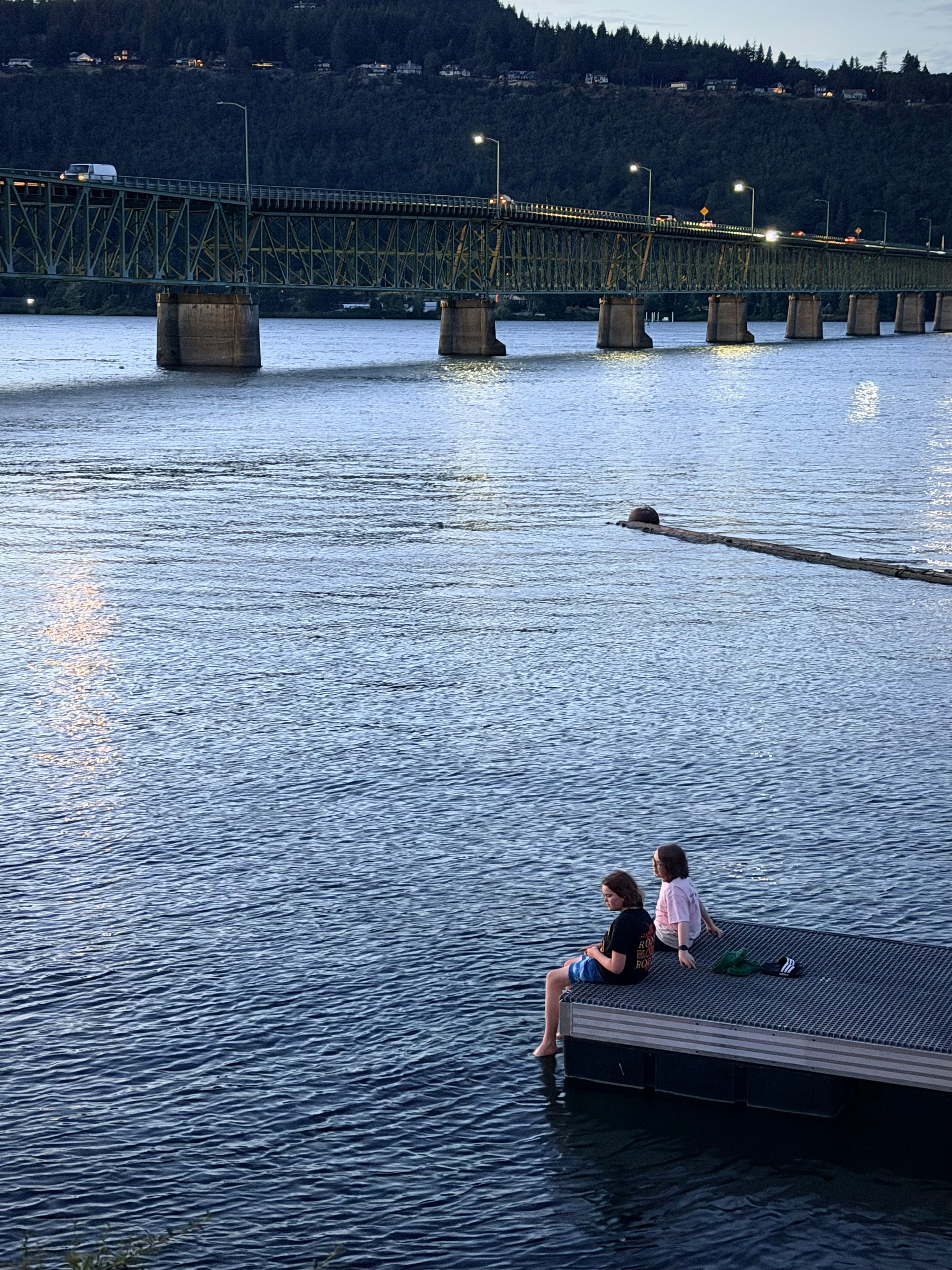 Two children sit side by side on the edge of a floating dock with their feet in the Columbia River as dusk settles in. The Hood River Bridge spans the background while calm water fills most of the frame.