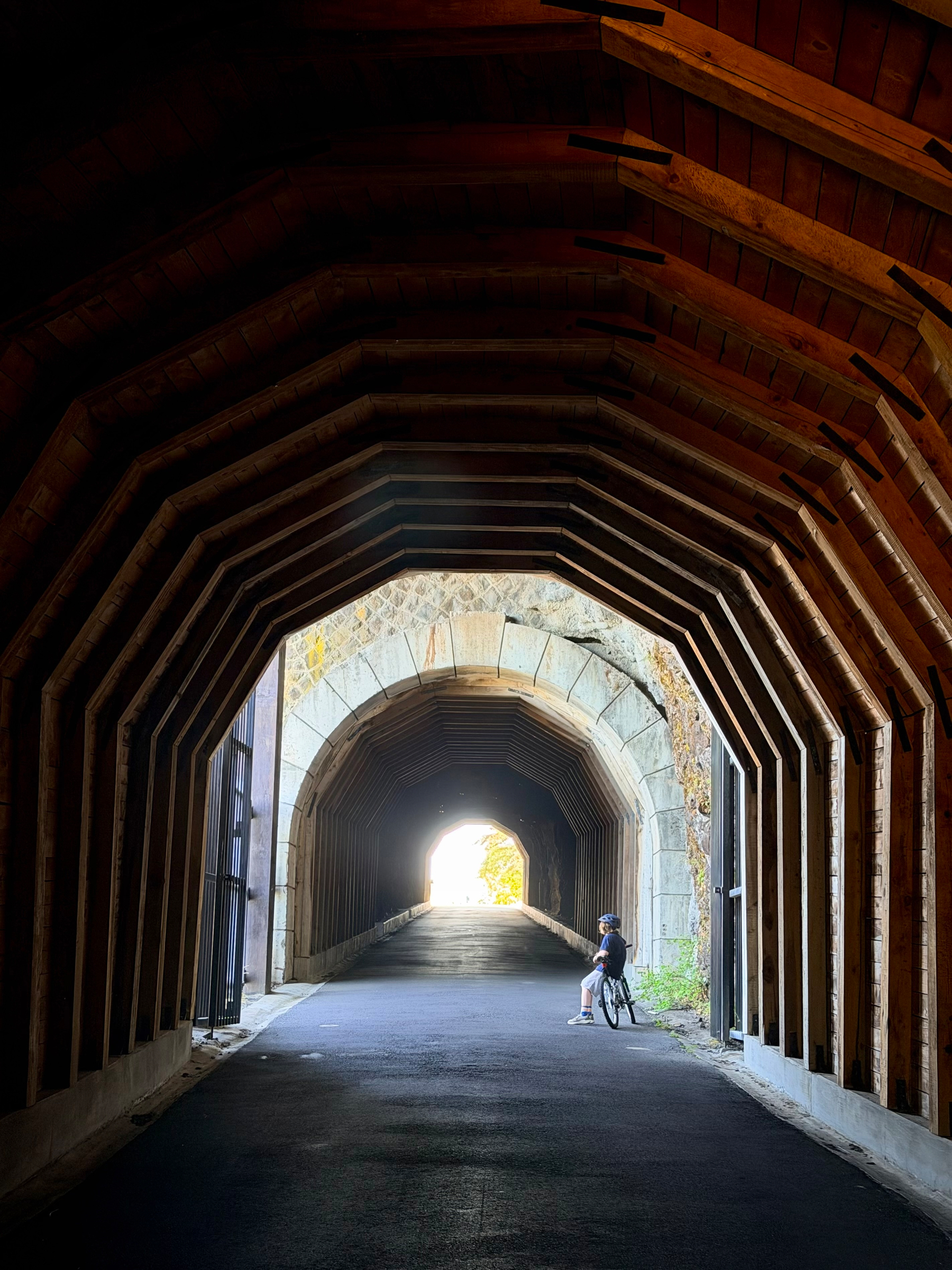 A child pauses on a bicycle inside a timber-lined tunnel, silhouetted against the bright daylight at the far end. Repeating wooden beams frame the scene, creating a strong sense of depth and symmetry.