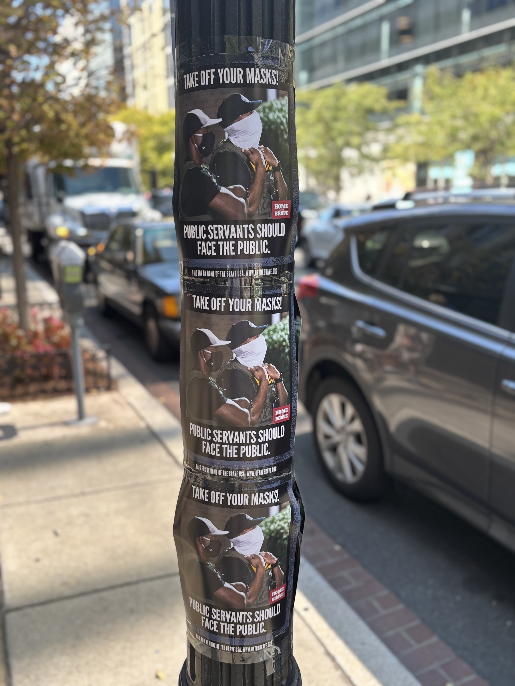 Posters on a street pole feature a person in a mask with the message "TAKE OFF YOUR MASKS! PUBLIC SERVANTS SHOULD FACE THE PUBLIC."