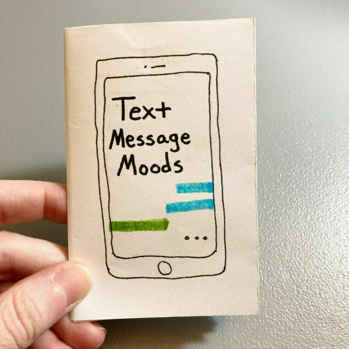 A hand holds a small booklet with a drawing of a smartphone on the cover, labeled Text Message Moods and featuring colored text bubbles.