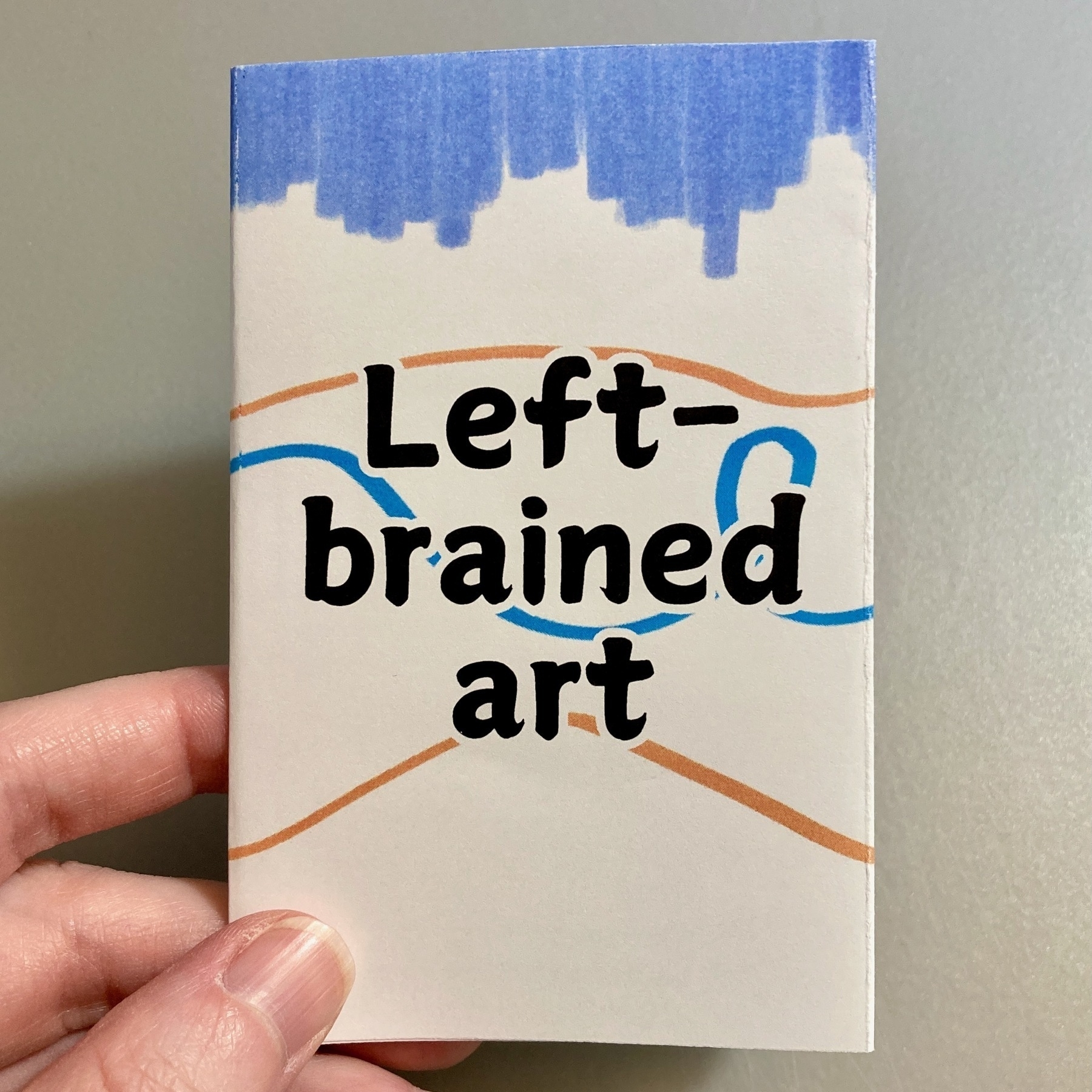 A hand holds a mini zine called Left-brained art.