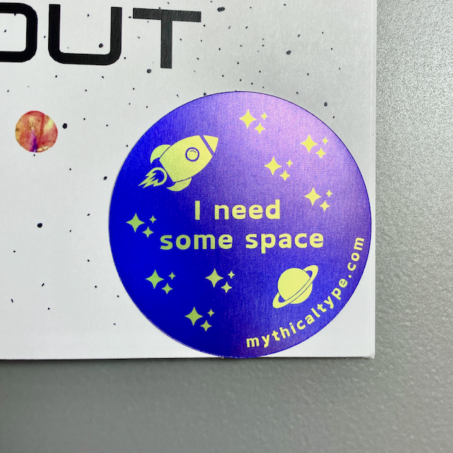 A round holographic sticker that says I need some space. Text and graphic elements, including a rocket ship, planet, and stars, are in yellow on a purple background.