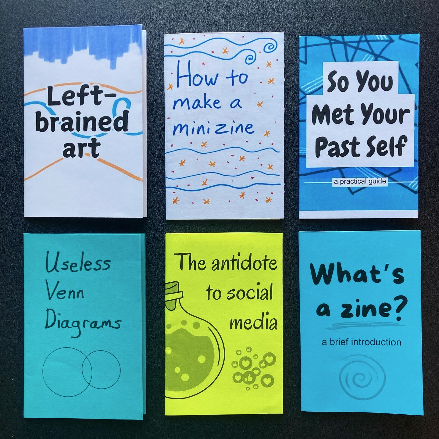 Six colorful mini zines with various titles such as Left-brained art and How to make a mini zine are displayed on a dark surface.