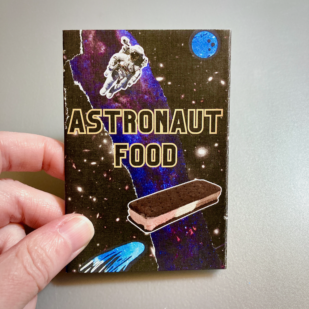 A hand holds a mini zine titled Astronaut Food. The background is a collage of outer space imagery. There’s an astronaut above the title, and a freeze-dried ice cream sandwich below the title.