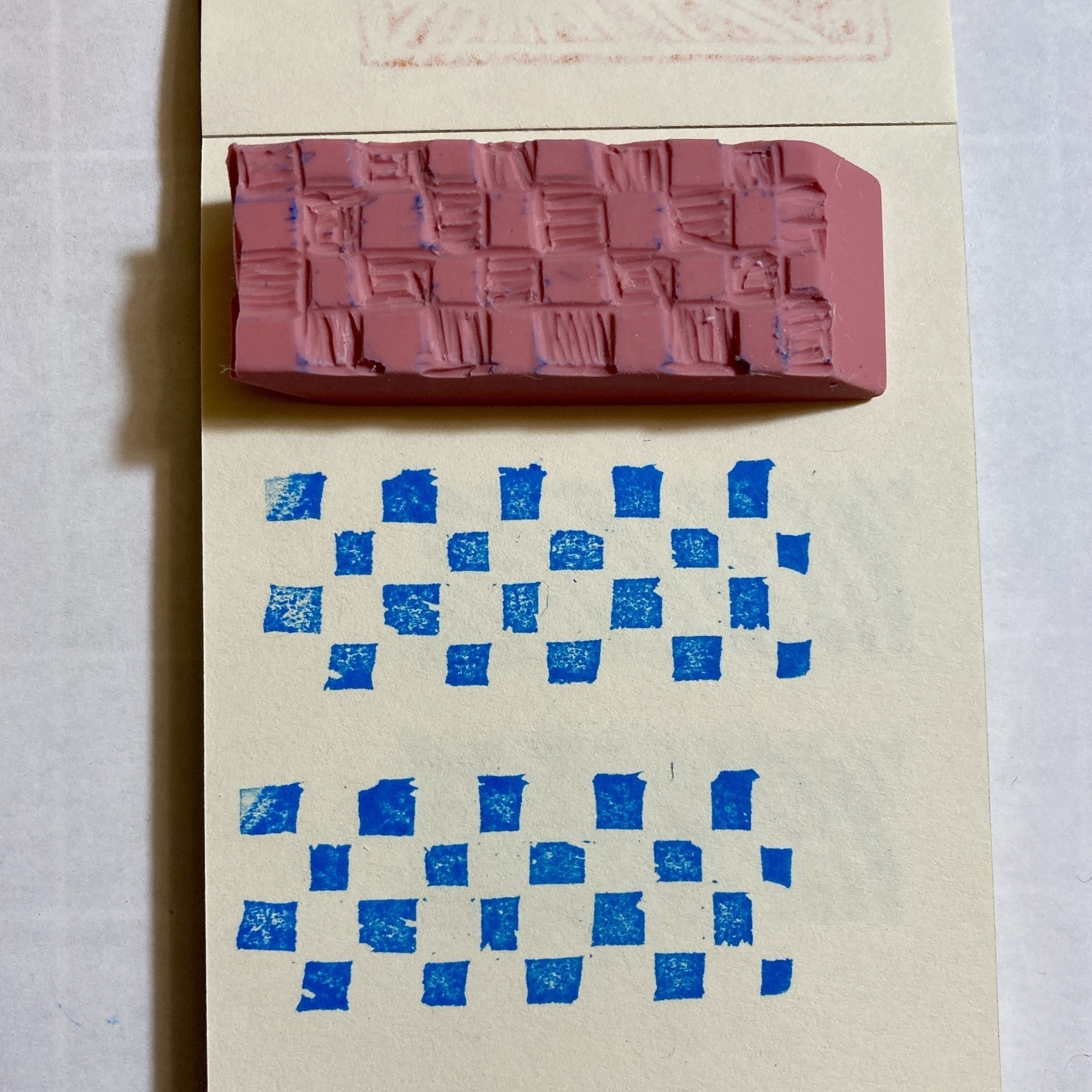 A carved pink rubber stamp on a piece of paper with two stamps showing a blue checkerboard pattern.