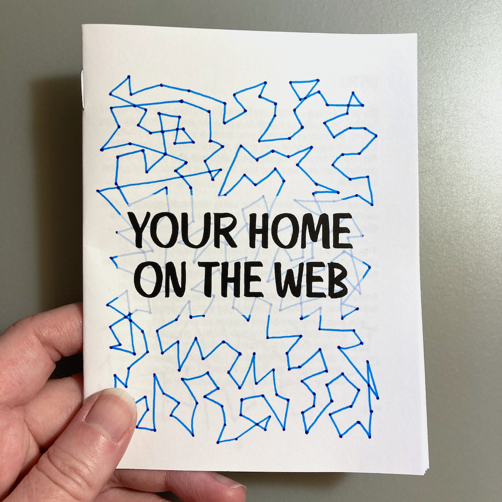 A hand is holding a zine with abstract blue line drawings and the text YOUR HOME ON THE WEB on the cover.