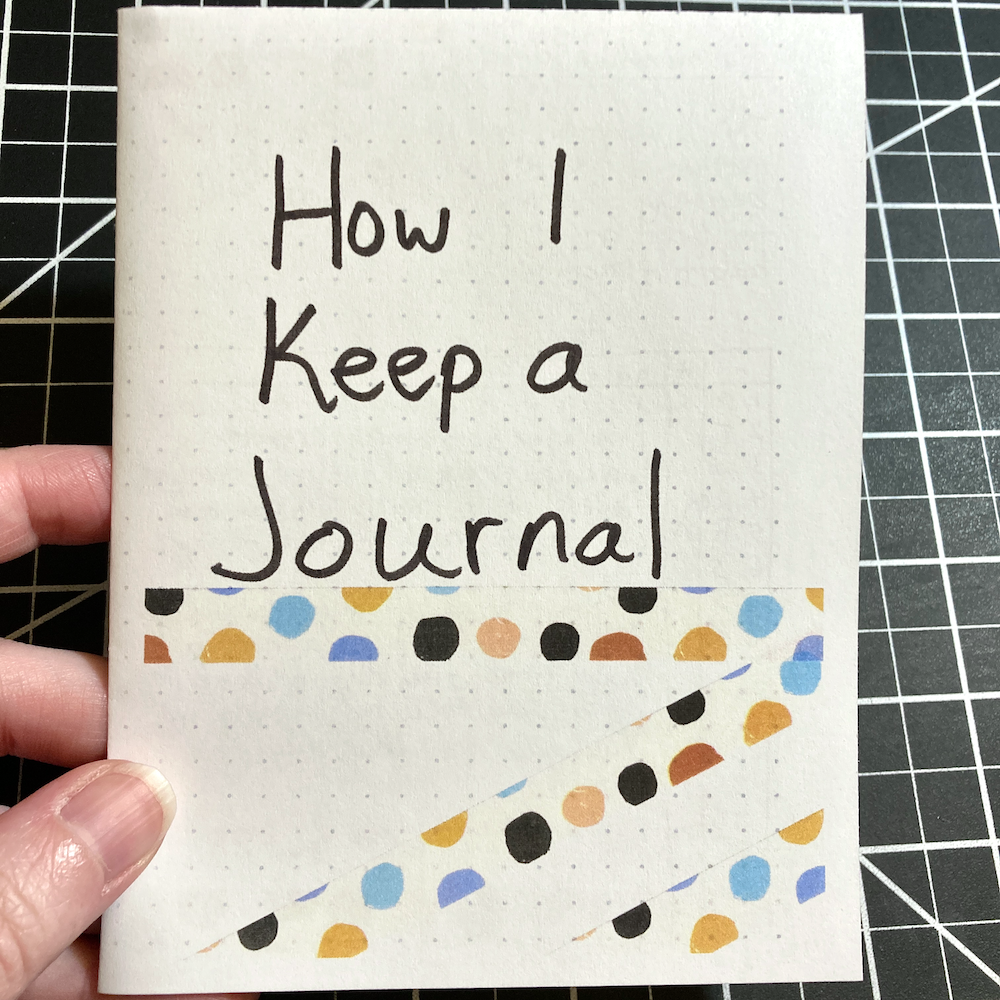 A hand is holding a zine with the title “How I Keep a Journal” written on the cover and decorated with colorful polka-dotted tape.