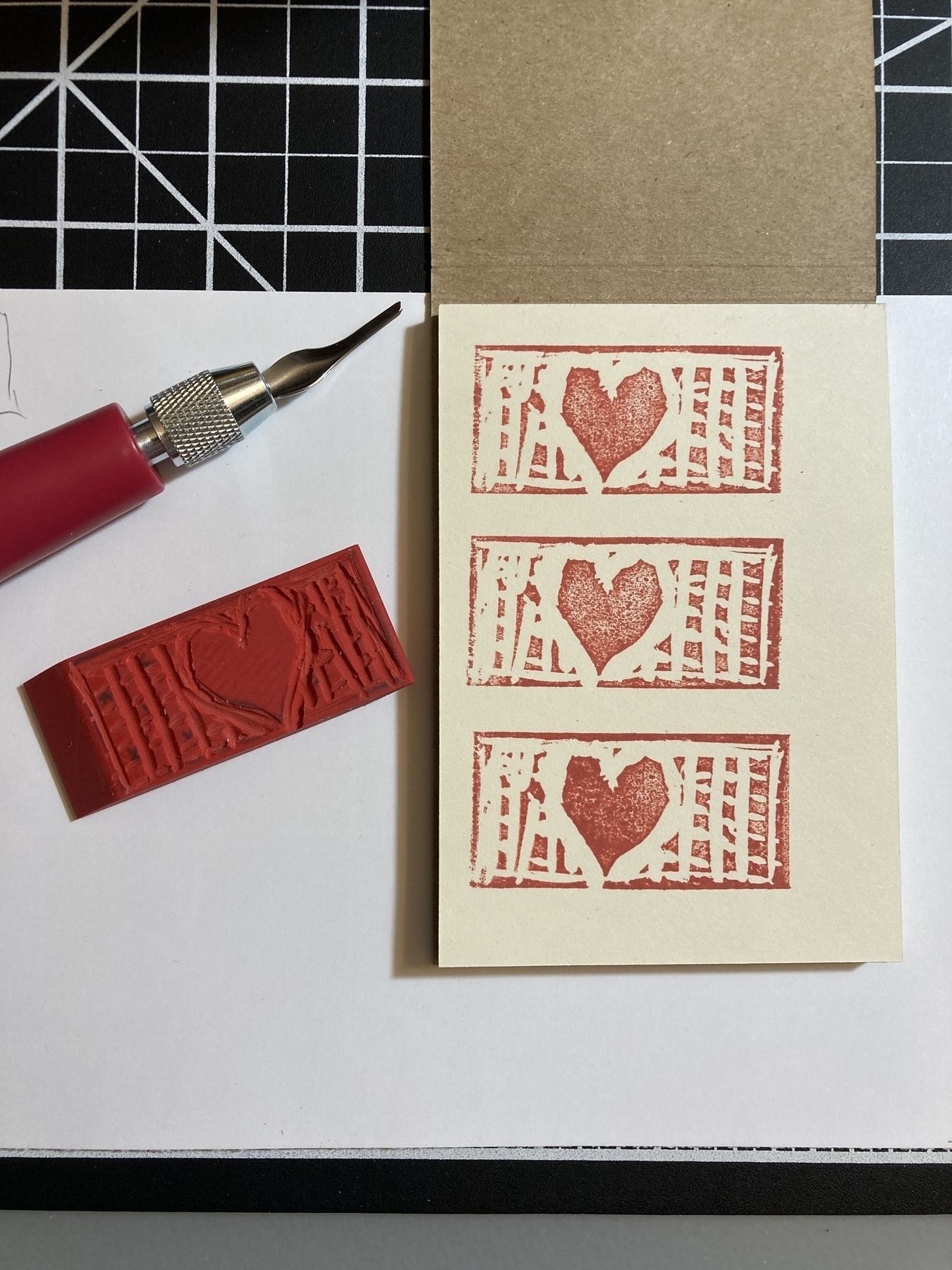 A hand-carved stamp of a heart design and cutting tool are positioned next to stamped heart designs on paper.