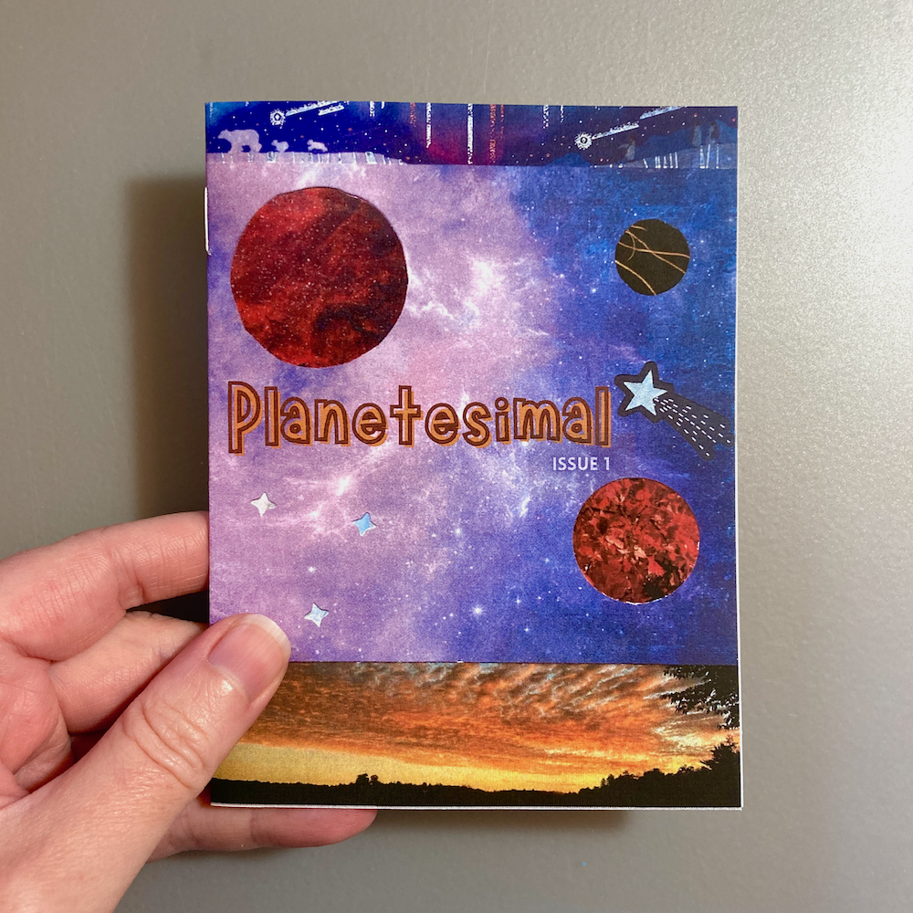 A hand holds a small zine titled Planetesimal Issue 1, featuring a cosmic-themed cover with planets and a sunset landscape.