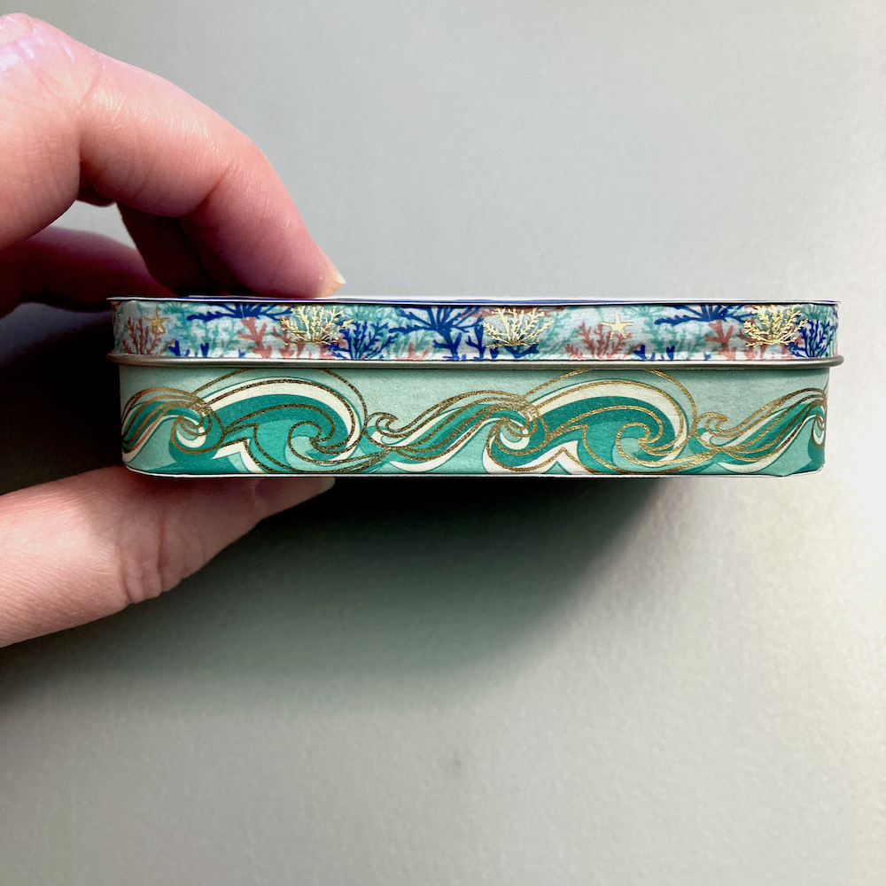 A hand holds a decorative, illustrated tin box with an ocean-themed design.