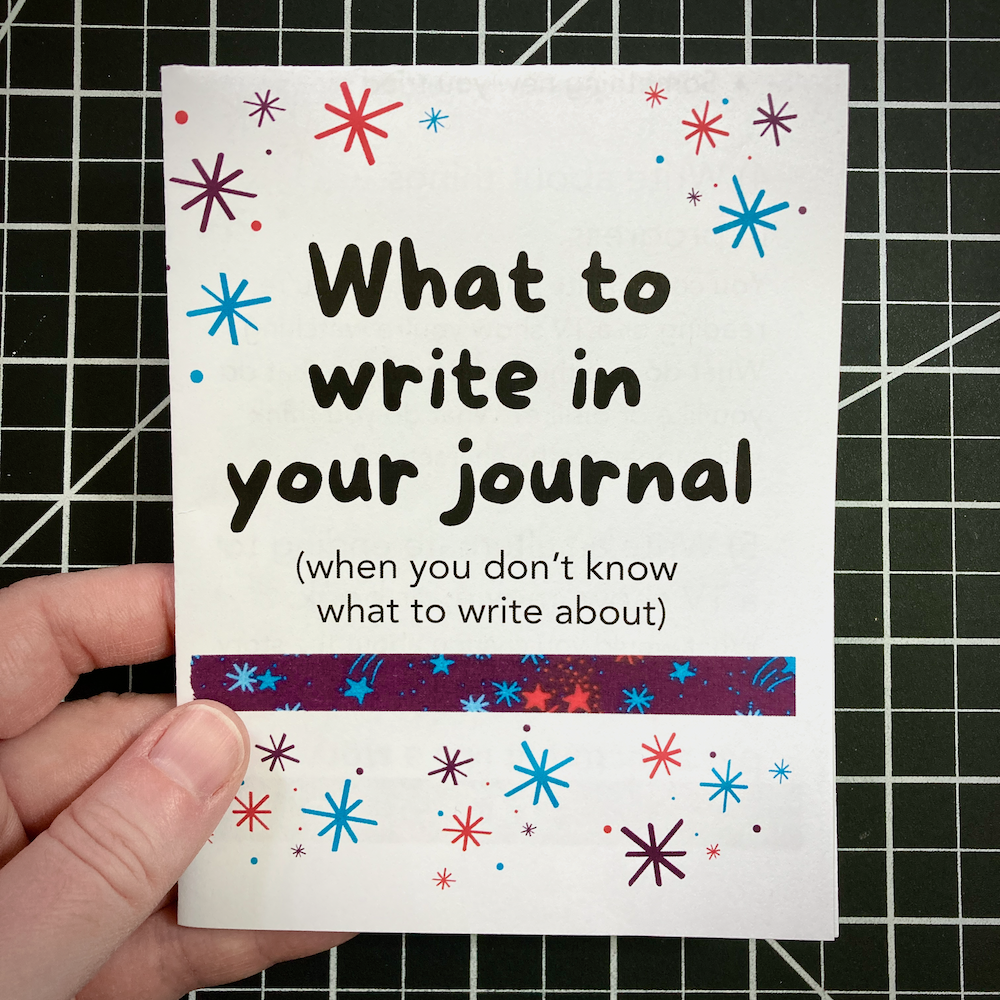 A person holds a zine titled What to write in your journal (when you don&rsquo;t know what to write about) against a grid background.