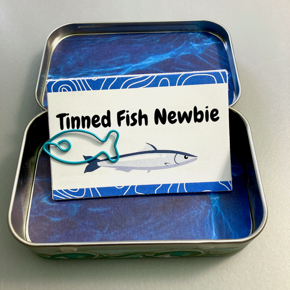 An open tin box contains a tiny zine titled Tinned Fish Newbie with a fish-shaped paperclip attached.