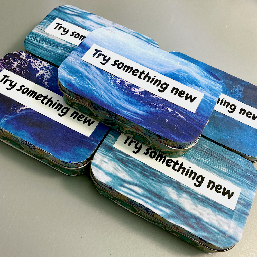 Several rectangular tins with ocean wave designs and the phrase Try something new printed on them are stacked on a table.