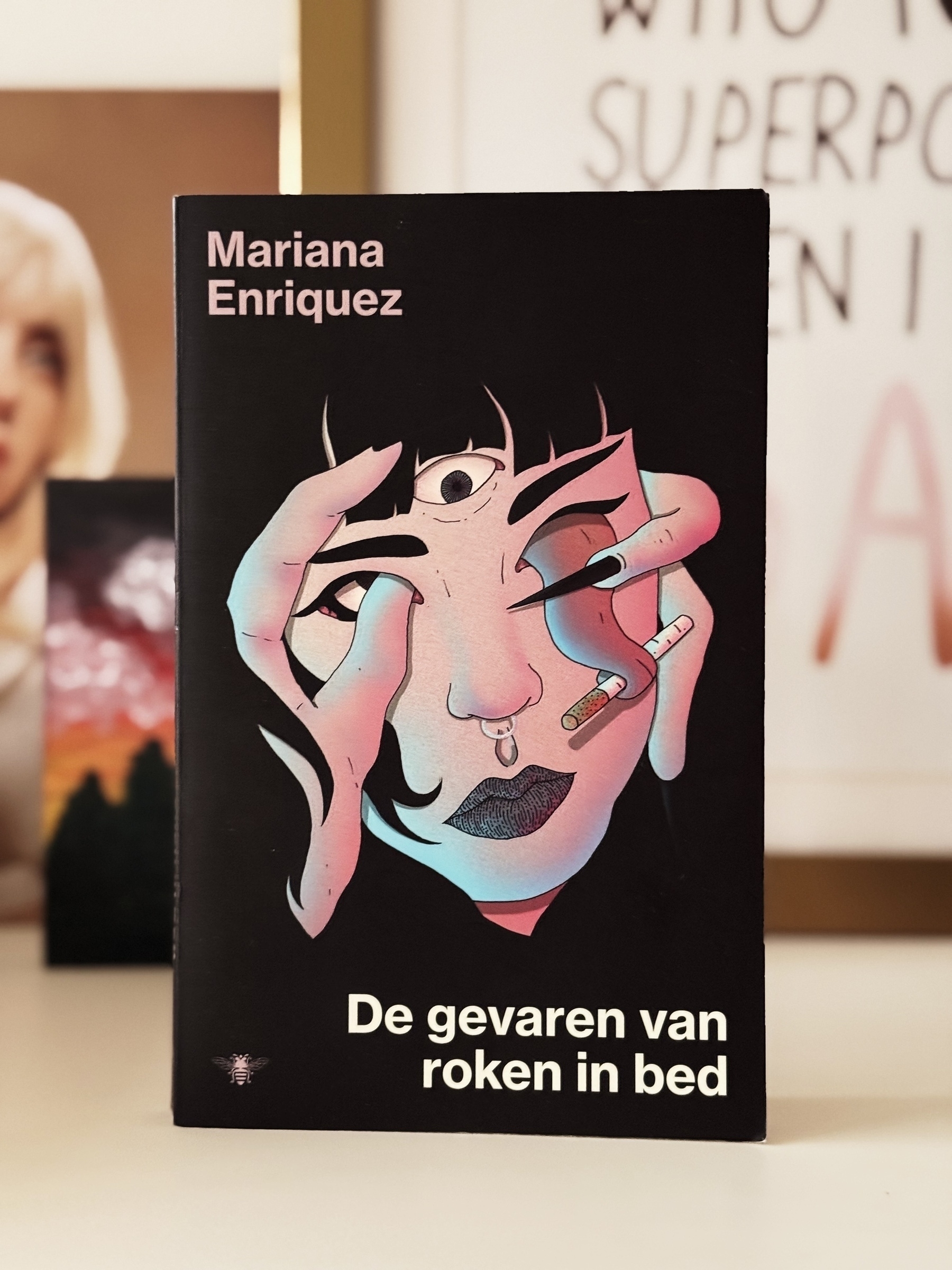 A book titled De gevaren van roken in bed by Mariana Enriquez features an abstract illustration of a face with hands, eyes, and cigarettes.