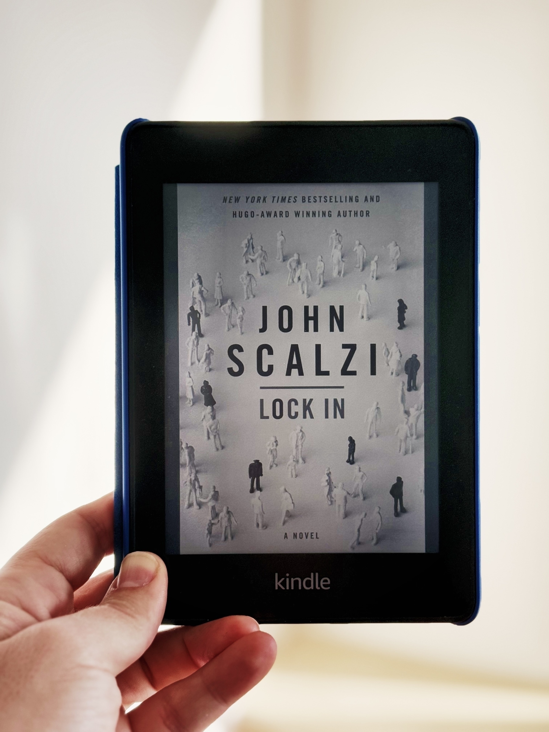 A person is holding a Kindle displaying the cover of the book Lock In by John Scalzi.