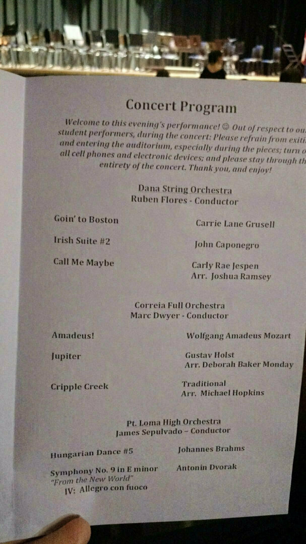 A concert program with a list of musical pieces and their respective conductors and performers is being held up in front of a stage with musical instruments.