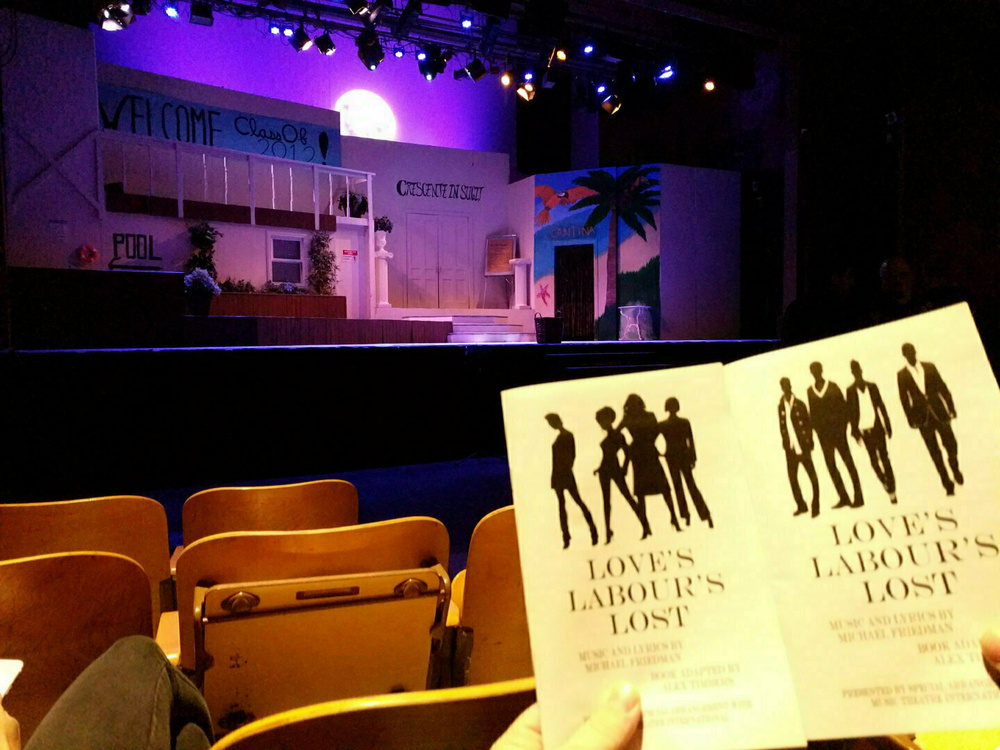 Two people are holding playbills for Love's Labour's Lost in a theater with a stage set for a performance.