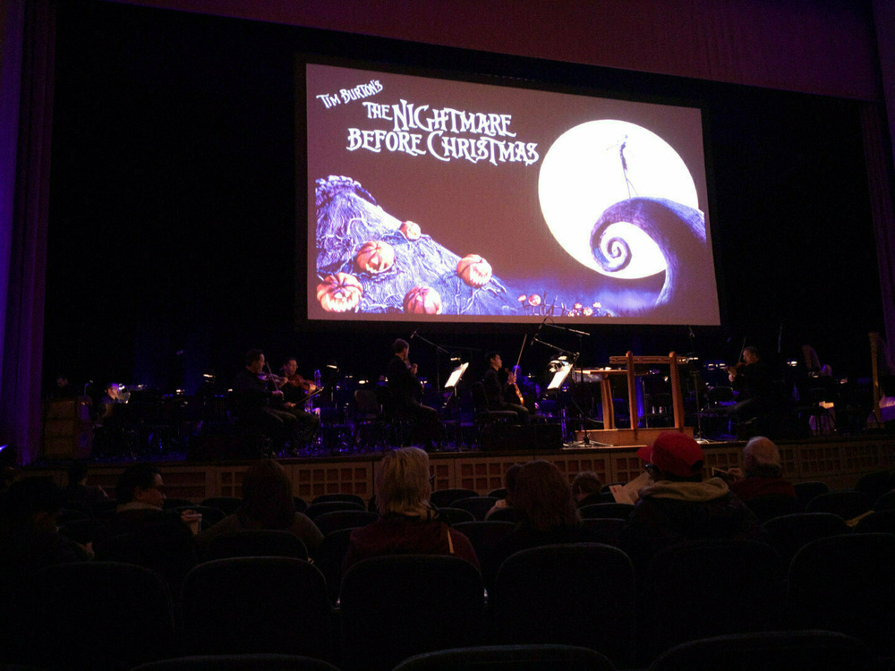People are seated in a theater with a large screen displaying Tim Burton's The Nightmare Before Christmas above an orchestra.