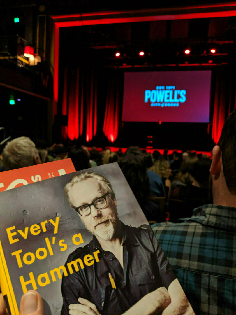 An audience in a dimly lit theater is preparing for an event, with a person holding a book titled Every Tool's a Hammer in the foreground.