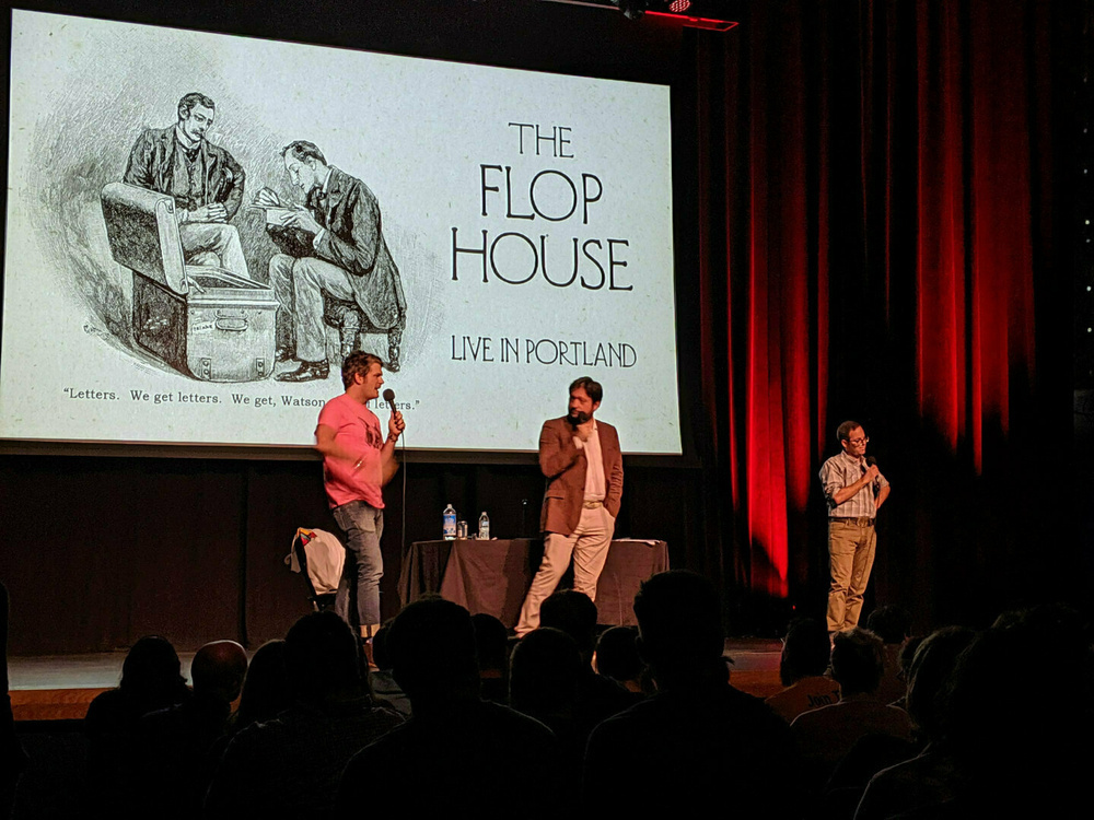 Three people are on stage hosting a live show in front of an audience, with a screen displaying The Flop House Live in Portland.